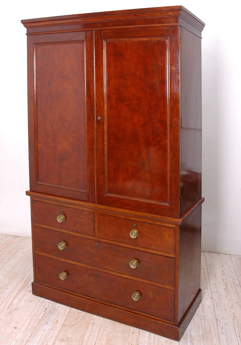 In figured African mahogany, this English linen press offers four interior tray drawers behind two cabinet doors with reversed hand-chamfered panels. The bottom half offers two small drawers over two long drawers and rests on a plinth