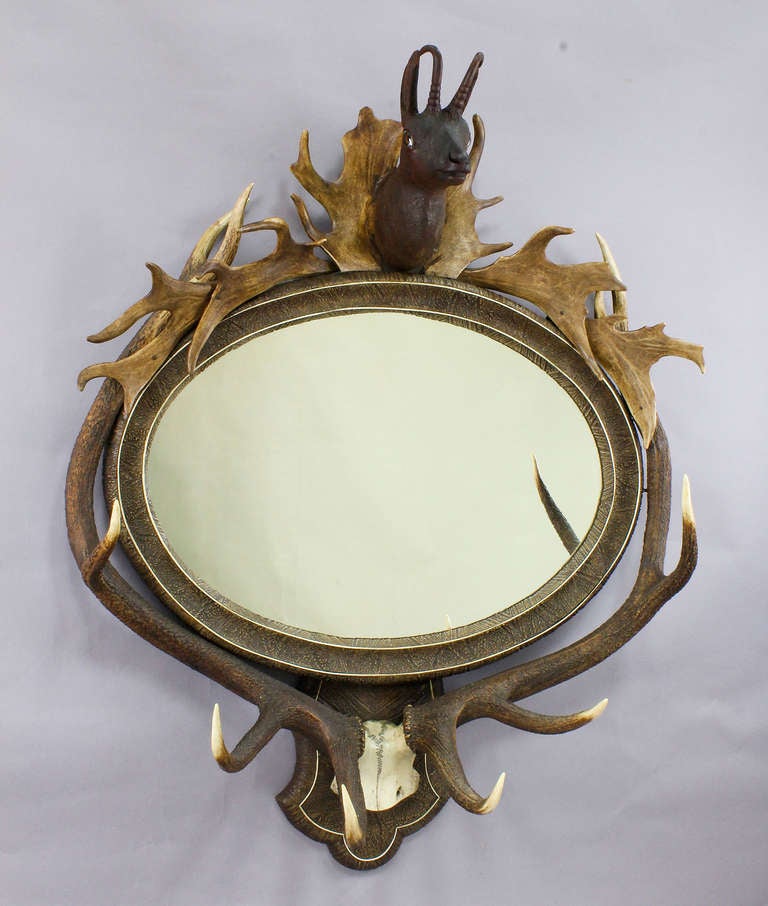 a large oval antler mirror, austria ca. 1860. frame decorated with a large deer trophy, fallow deer antlers and a wooden carved chamois head on top. mirror frame covered with stucco imitating an antler veneer.