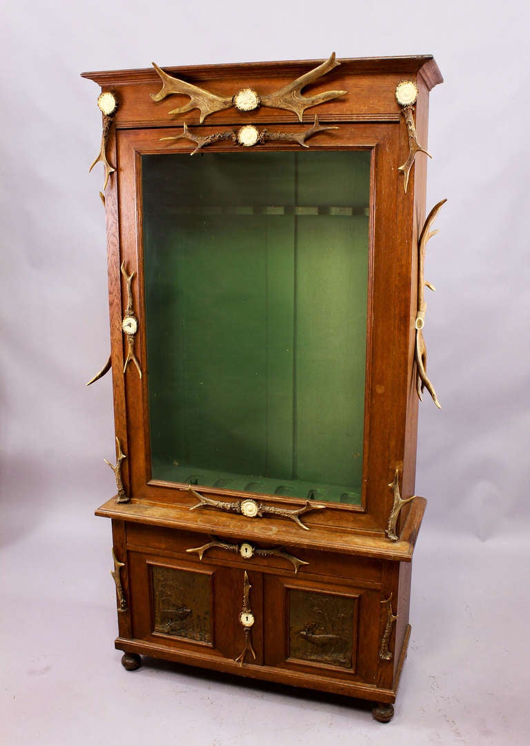 Antique wooden gun cabinet decorated with original antlers from the deer, fallow deer and turned horn roses. With glass door, the lower door panels are metal plates depicting stags. For six rifles.