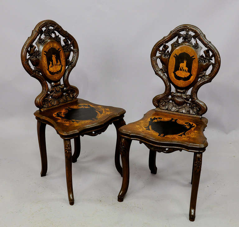 a wonderful pair of carved walnut chairs with finely inlaid seat and back. superb carved edelweiss and floral ornaments. executed ca. 1900, swiss brienz. (the center paintings at the seating surface overpainted in black)