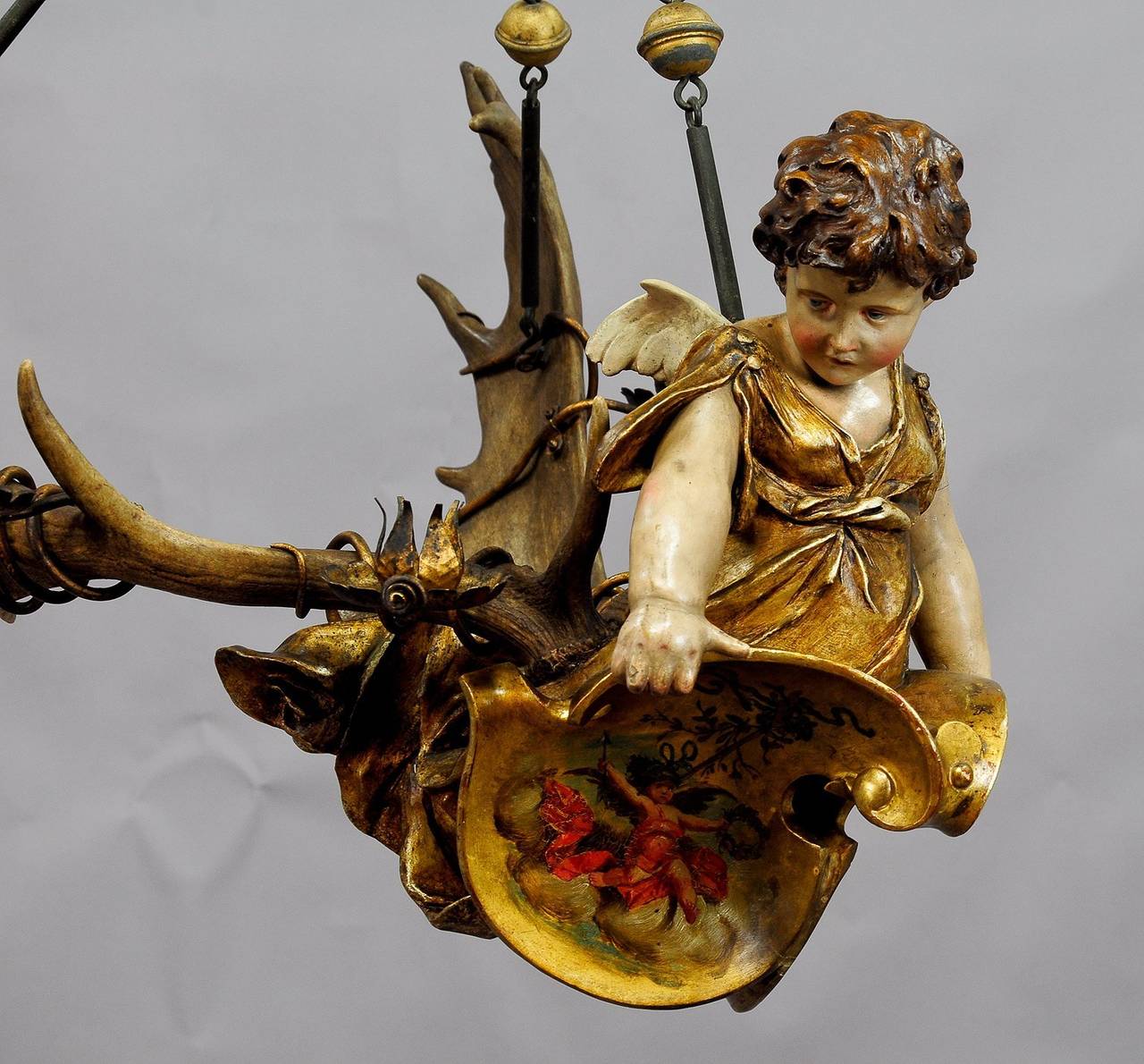 an antique statue of a cherub made of handpainted plaster - mounted on a pair of original fallow deer antlers. with 6 handforged and gilded iron candle holders. the angel is holding a pair of emblems with lovely angel paintings on them. executed ca.