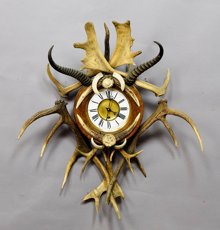 An antique black forest antler wall clock. Enamelled clock face with painted figures. Decorated with original antlers from the deer, fallow deer and mountain goat, wild boar tusks and turned and carved horn ends. One antler with brass emblem - 