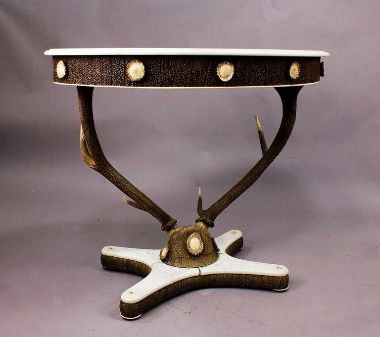 a sculptural antler table, austria ca. 1860. a large stag trophy is holding the table top, base and table top with marble slap. rim of base and table top covered with stucco cover imitating an antler veneer. literature reference: "Geweihmoebel,