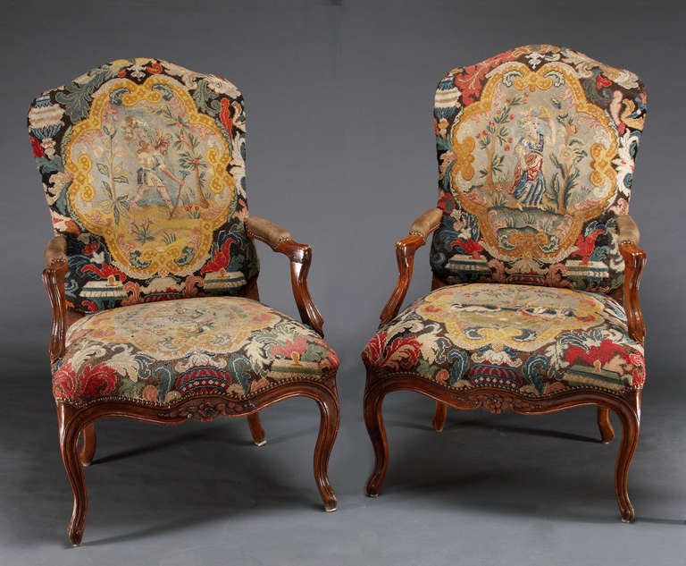 A pair of Louis XV Period walnut fauteuils, each with an arch back, serpentine seat rail with floral carving detail. Needlepoint appears to be of the period, with the figures of a male on one chair and a female on the other chair. Chairs are