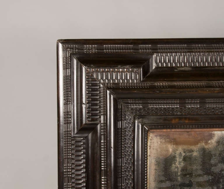 The original mercury mirror plate is inset into a compound rectilinear frame of ebonized fruitwood; with alternating carvings of rope, weave, and ripple ornamentation. Elegant dimensional character to intricately carved frame. Aging to mercury