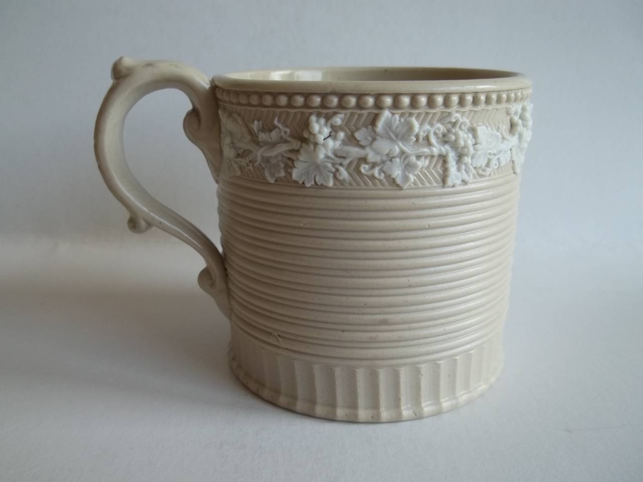 This is a large Coffee Can or Mug which I attribute to the Wedgwood factory.

The mug is cylindrical in form with a loop handle having upper and lower spurs. and shell-like moulded terminals, where the handle meets the body.The dark cream / stone