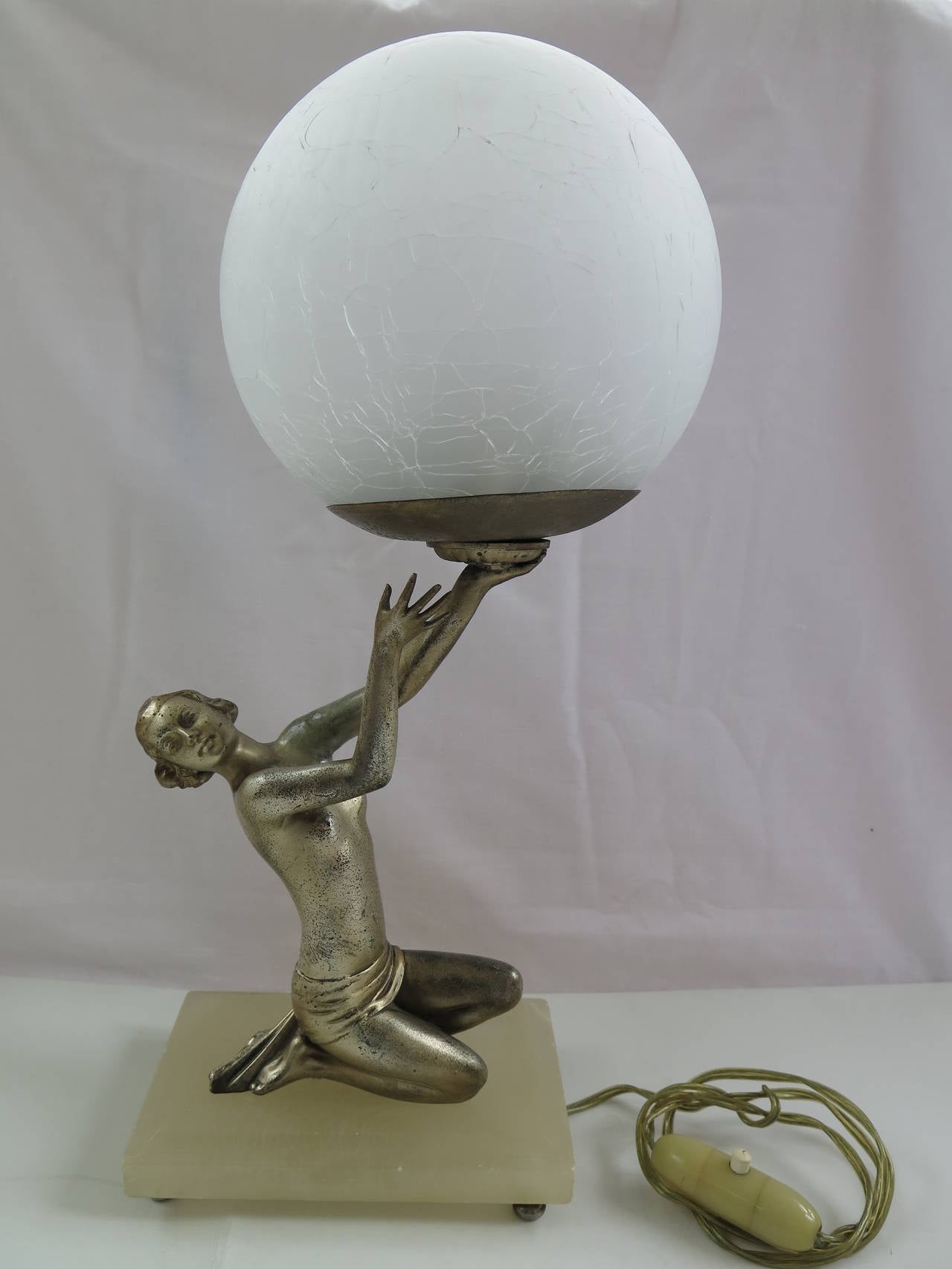 This is a very good table lamp, original to the Art Deco period.

The shelter lamp has a lady in a kneeling position holding the lamp above her head. The lady figurine is slim and scantily clad in just a skirt, typical of the stylized figures of