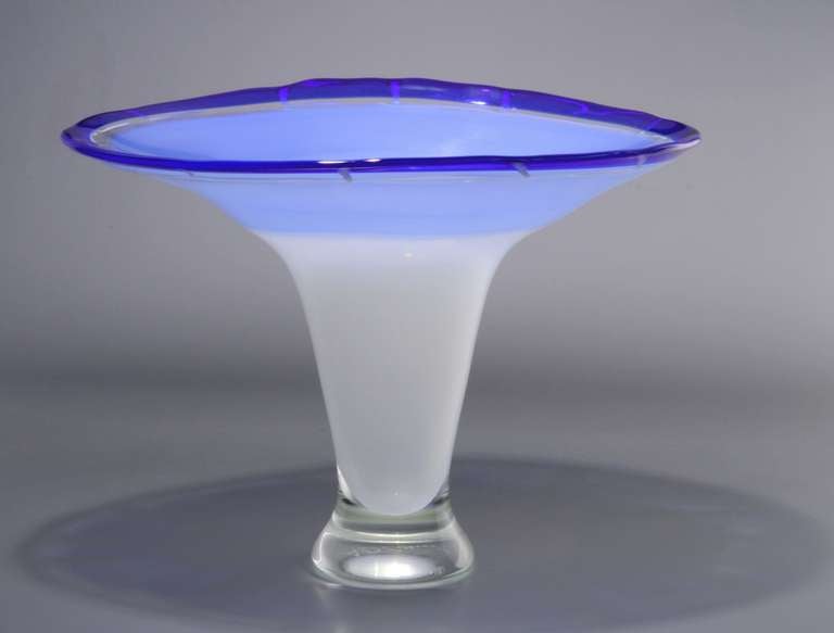 Elegant one-off vase in opaque white, light blue with a dark blue rim. The vase has a conical and flaring shape and rim with white tread applied to it at intervals. The shaft is also flaring at the foot (colorless glass).

This unique studio glass