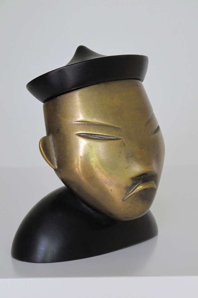 This stylized head of an Asian man, made of brass and blackened wood, is produced by the Viennese Hagenauer Werkstätte (Wien, Austria) around 1930. This rare piece is marked on the bottom (see photo). 

The Hagenauer Werkstätte focused on human