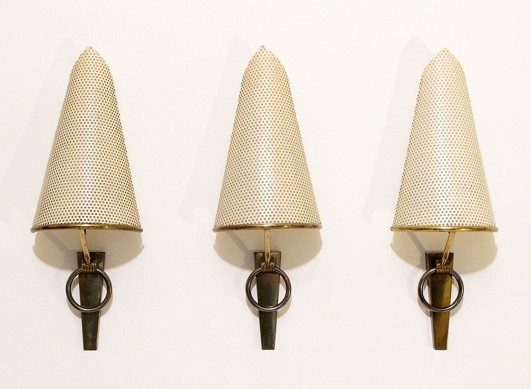 A set of three 1950s wall sconces by French designer Mathieu Matégot.

A fantastic modernist design, a soft, warm light is emitted from the perforations and reflected from the wall.

The angular cream-colored shades are held upon stylish brass