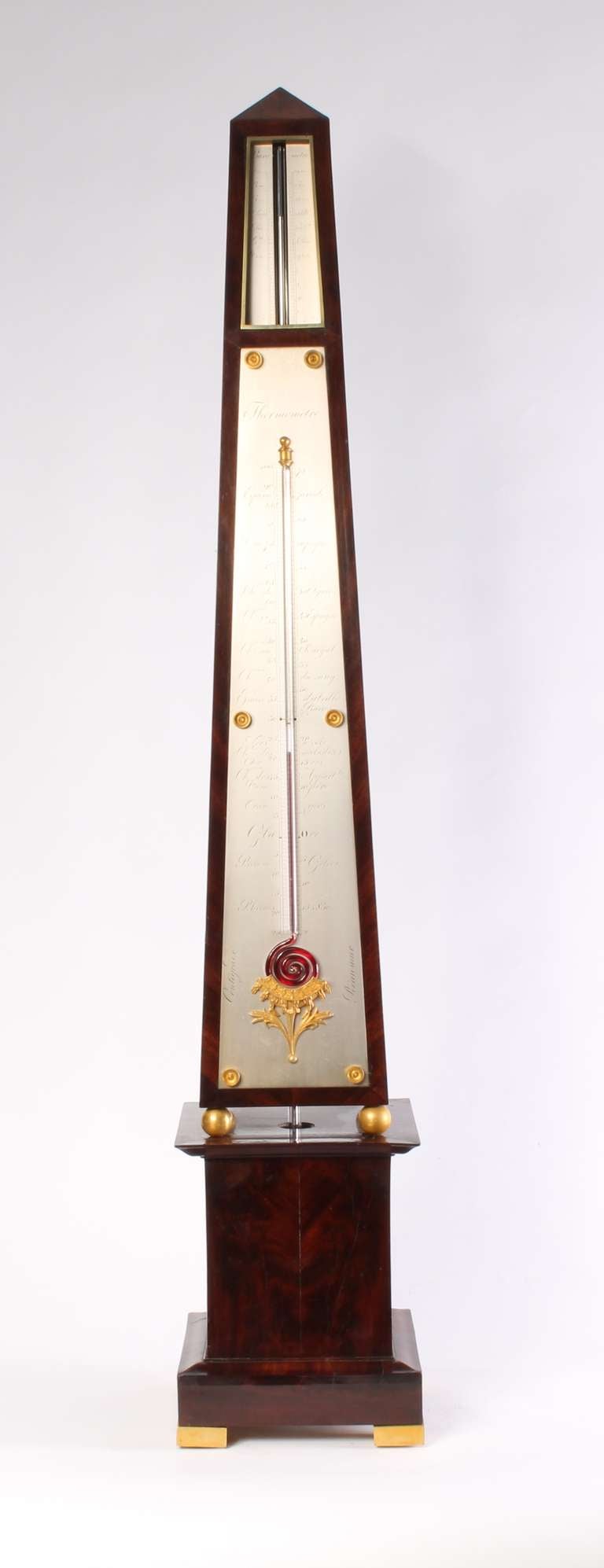 The flame mahogany-veneered pine case consists of two parts. The obelisk-shaped top has firegilt brass ball feet and is placed on a moulded base with firegilt brass block feet. The silvered brass register plates of the barometer are let in flush