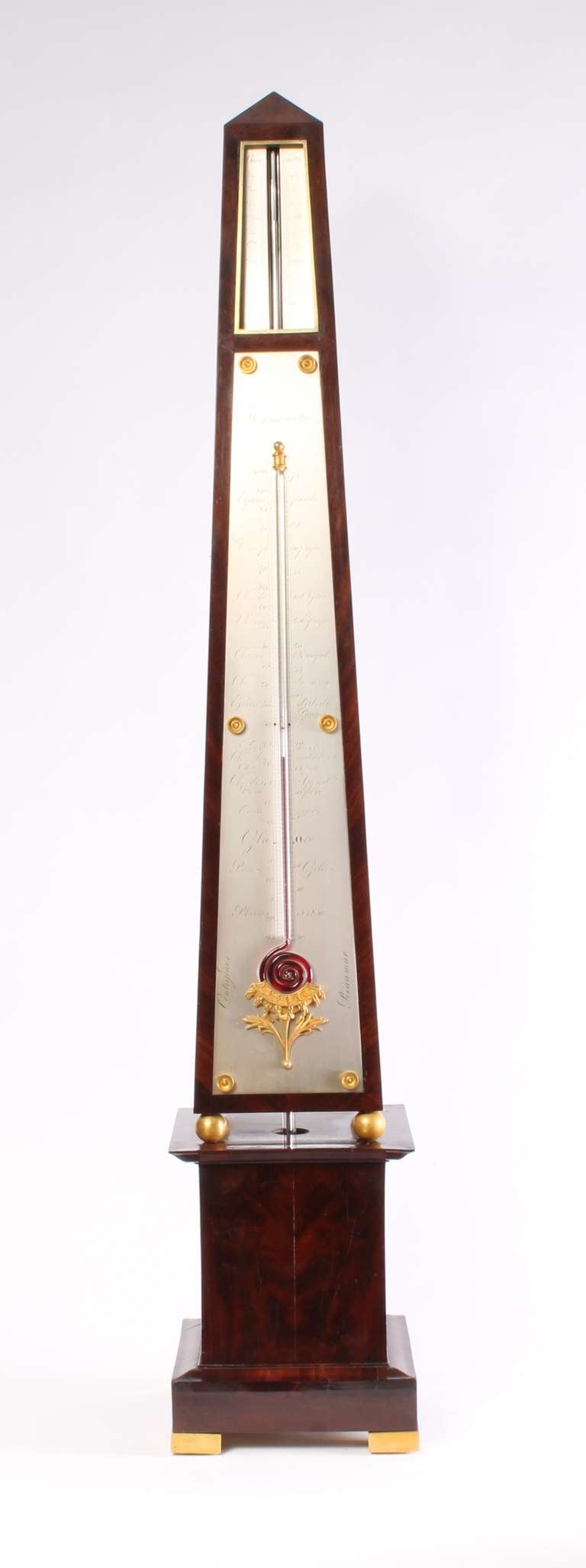 The flame mahogany-veneered pine case consists of two parts. The obelisk-shaped top has firegilt brass ball feet and is placed on a moulded base with firegilt brass block feet. The silvered brass register plates of the barometer are let in flush