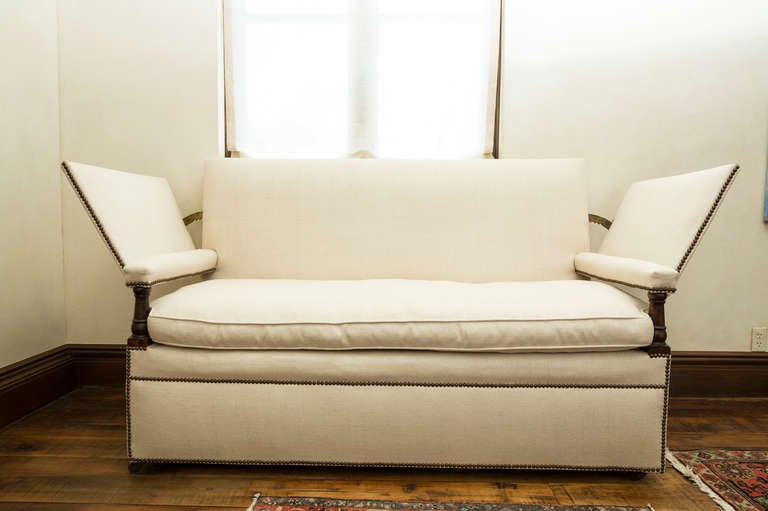 English ratchet arm sofa covered in white linen fabric with a single cushion. The sofa has wood columns on the arms.  The base has brass wheels with nail head trim continuing all the way around the sofa. The arms are working ratchet arms that