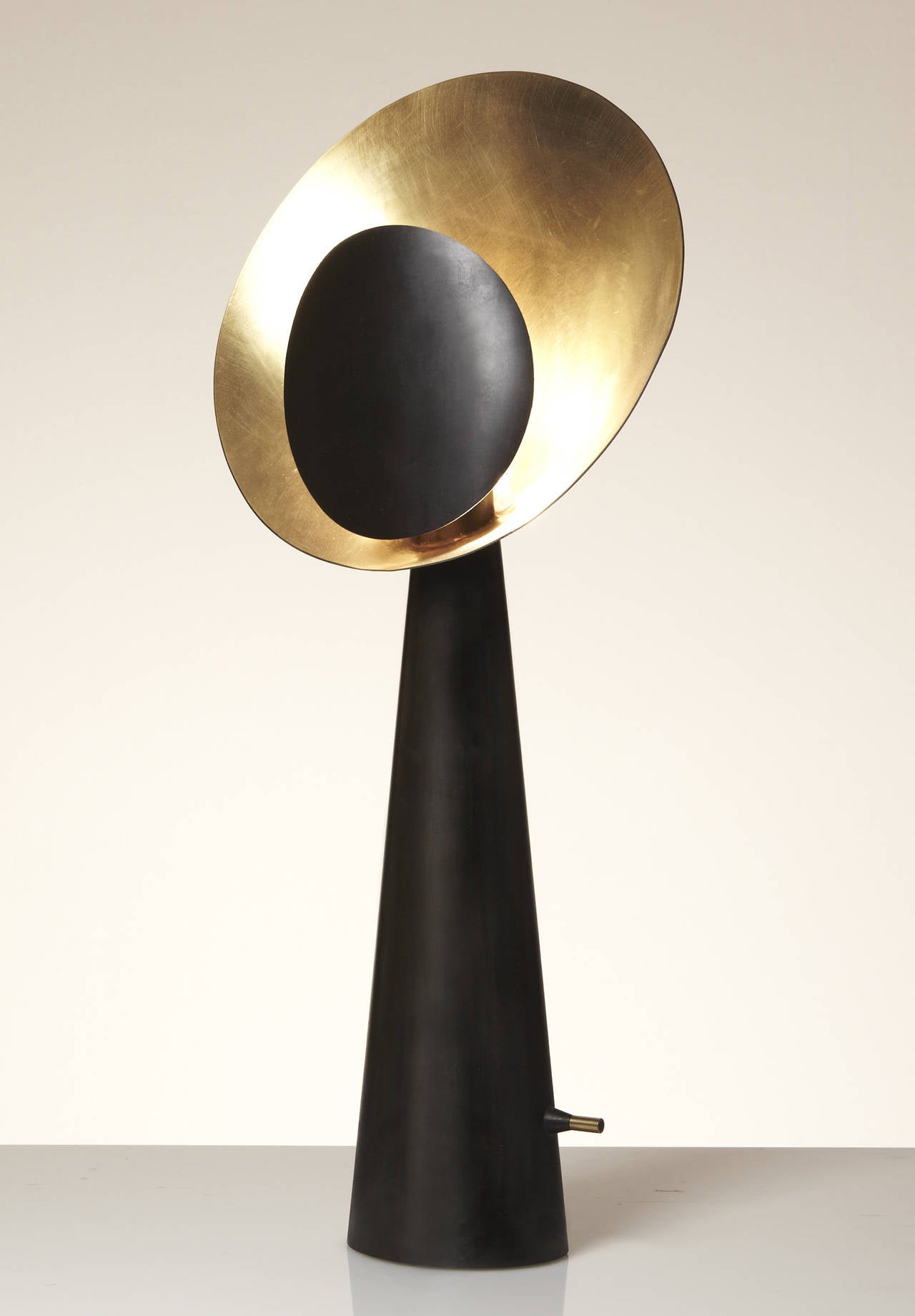 A sculptural hand-crafted half blackened table lamp made in gilded, polished and burned brass. 
The reflector shade optimize an irradiant ornamental light in warm golden tons. 
The light dimmer provides soft and diffuse light, or a bright light at
