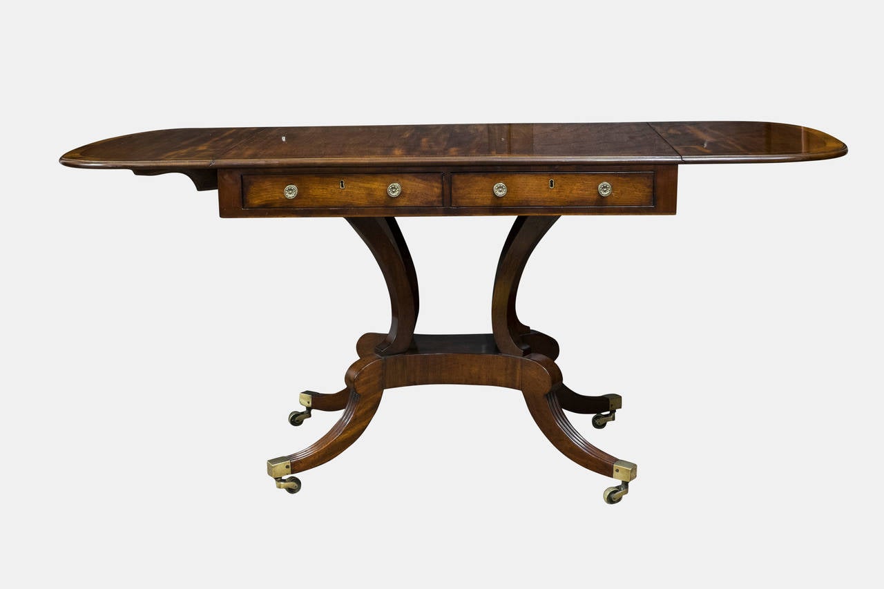 A Regency mahogany sofa table with rosewood crossbanding, two drawers with brass knobs and two opposing dummy drawers, a cluster base and swept reeded legs ending in brass casters, in excellent condition.