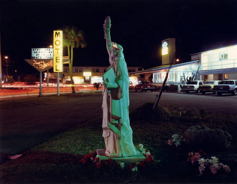 Sea Missle Motel, Cocoa Beach, Florida von David Graham ist ein C-Print im Format 30 x 40 Zoll, erhältlich in einer Auflage von 25 Stück. Dieses Foto zeigt eine kleine Freiheitsstatue auf einem nächtlichen Parkplatz, umgeben von Blumen. Dieses Foto