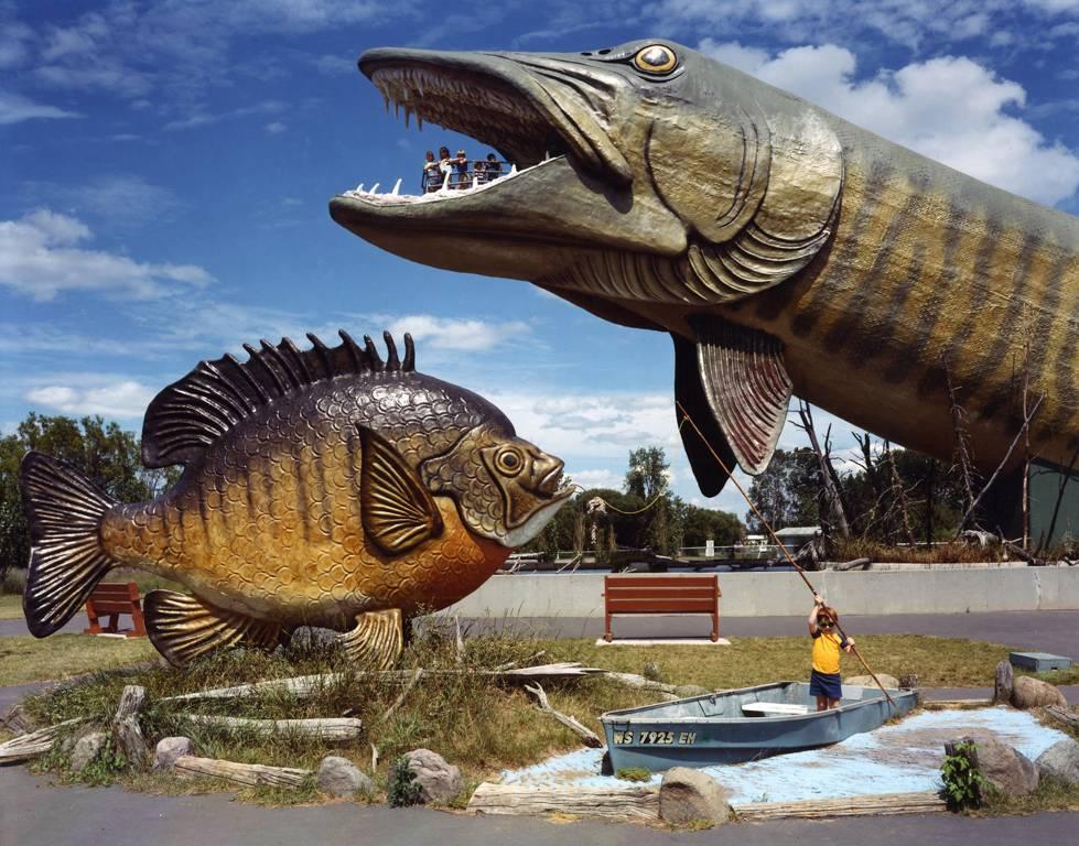 National Freshwater Fishing Hall of Fame, Hayward, Wisconsin par David Graham est une impression couleur de 20 x 24 pouces. Cette photographie représente deux faux poissons géants, avec un enfant debout dans un petit bateau et un groupe de personnes