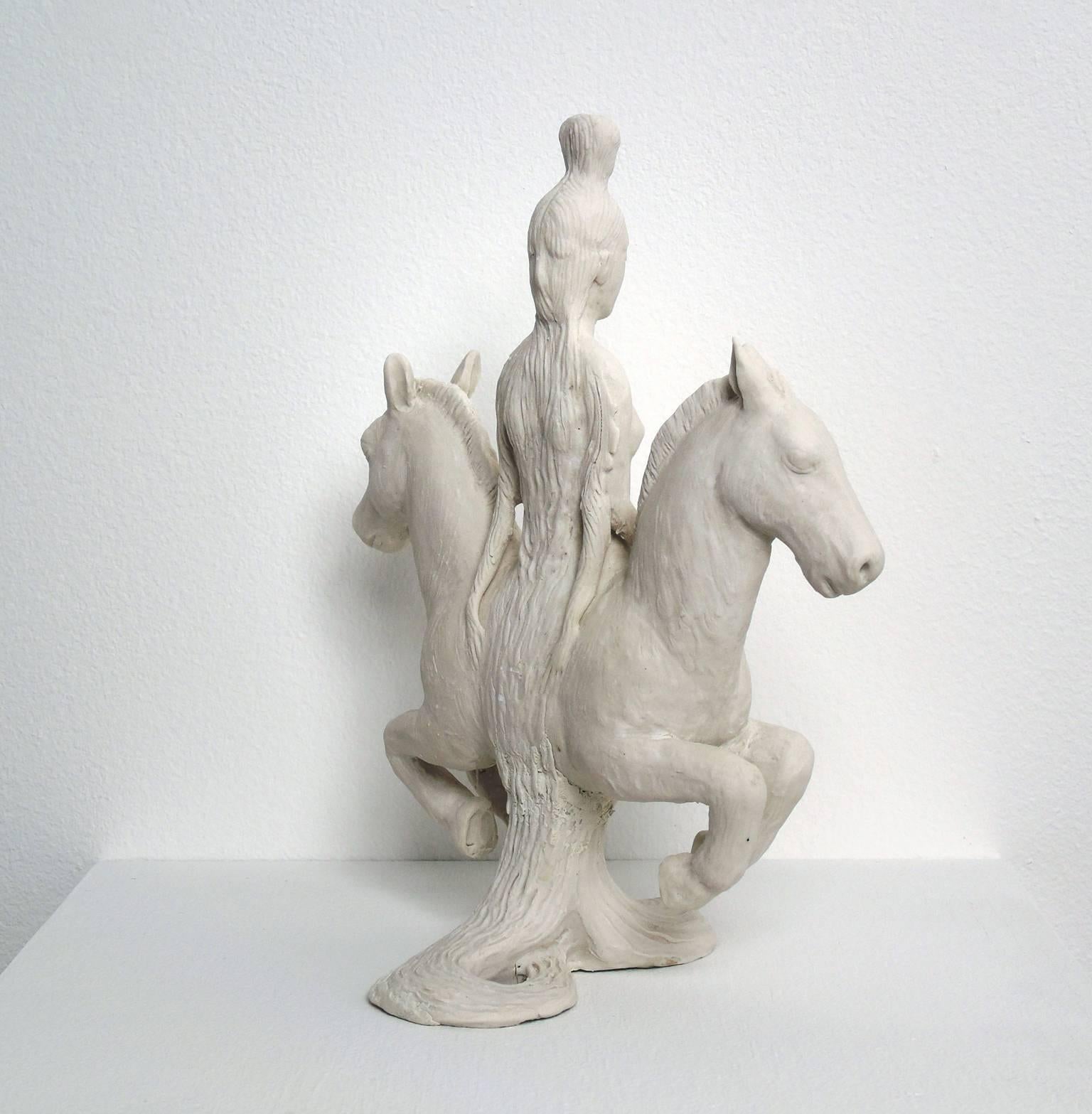 GODDESS, a white porcelain sculpture by Robin Whiteman, features a female torso centered on the back of a two headed donkey. The two donkey heads are at opposite ends of the beast. A pair of small faces emerge from the top of the girl's head, small