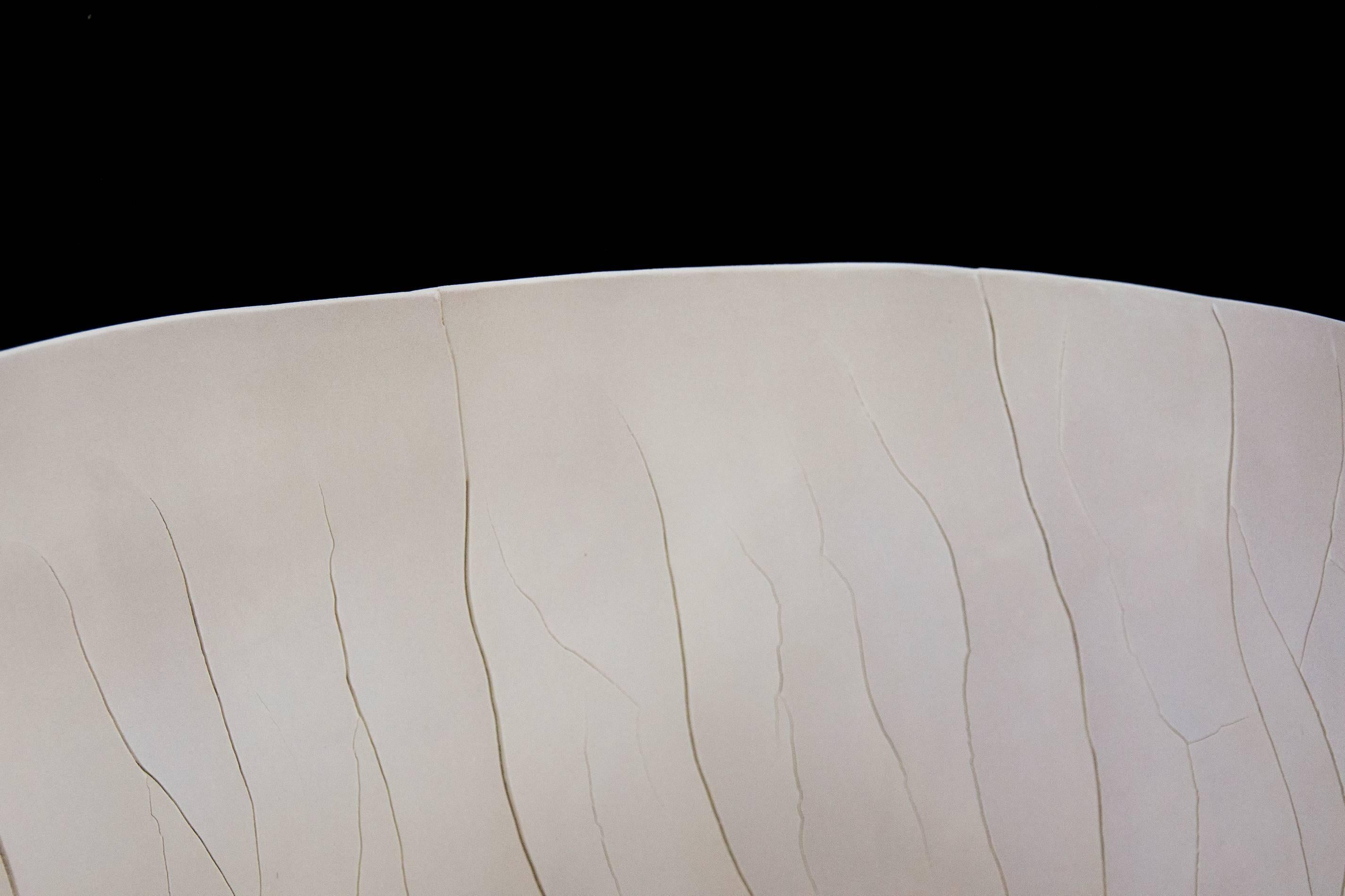 The polished surface of this off white 16 inch tall vessel is articulated with fissures created through the interaction of fibreglass filaments inserted into the wet porcelain clay before firing. The materials, color and texture of this piece speak