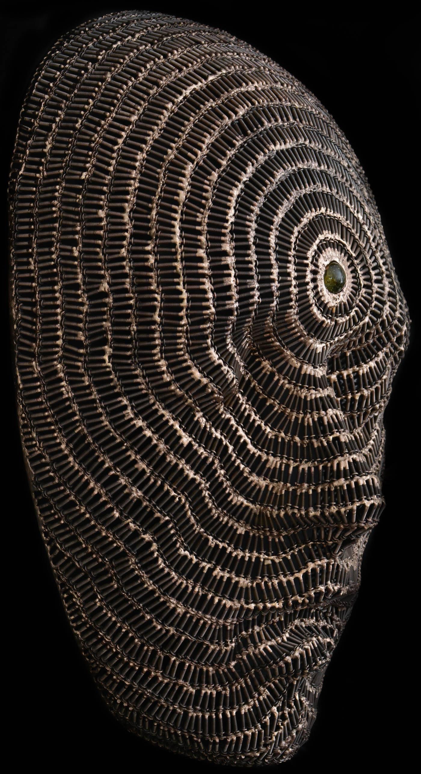 Rows of polished screws define the contours of a large face merging into concentric circles around a glass stone at the forehead. The title of this contemplative bronze and steel wall sculpture refers to the third eye or 6th chakra in the Hindu
