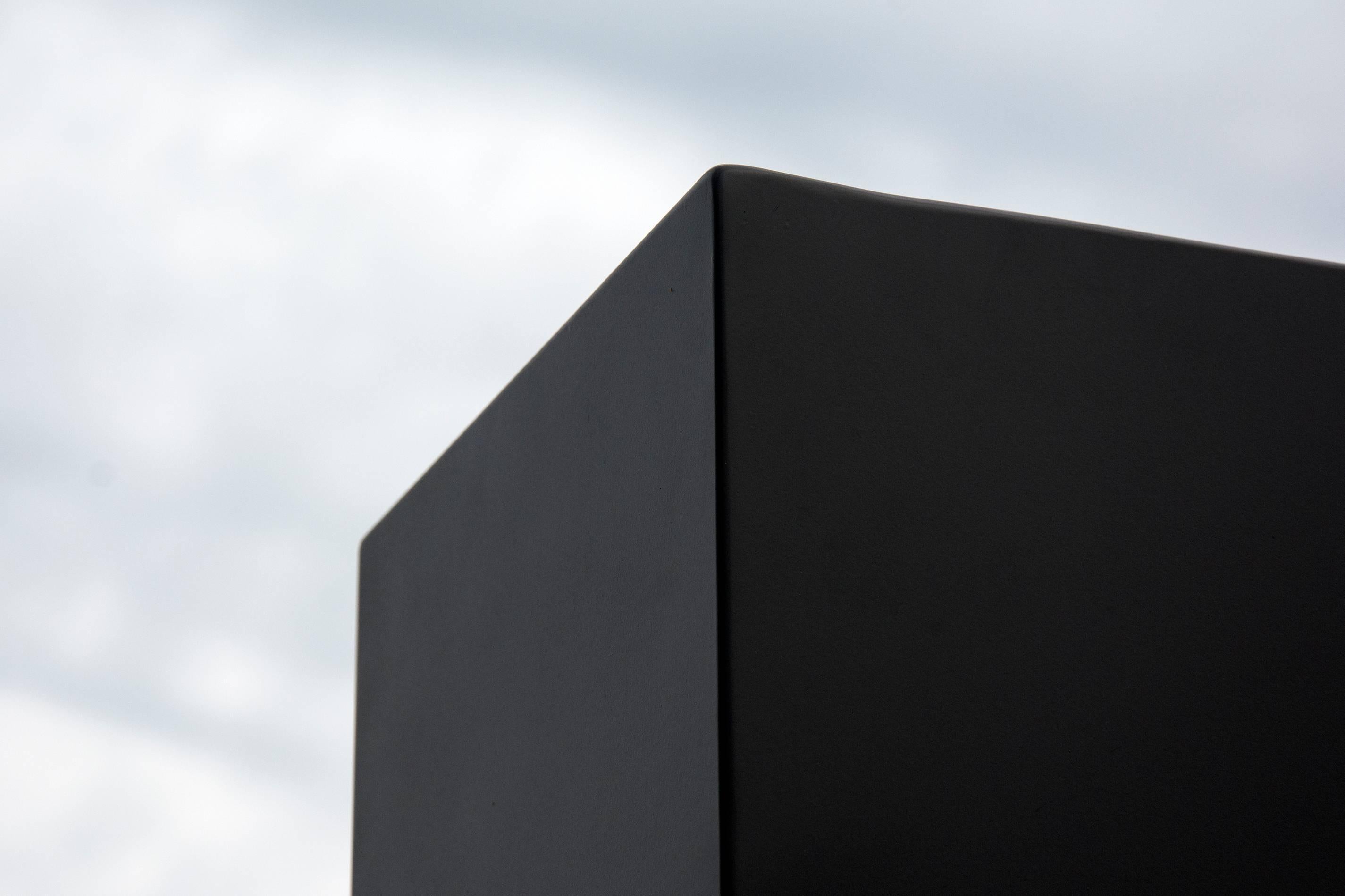 This piece is titled 'Aquagraphie' in French which translates to 'Water Writing'. A rectangular, steel outdoor sculpture in black is divided into three parts, like a puzzle, the edges polished and reflective. This seven-foot upright is mirrored in