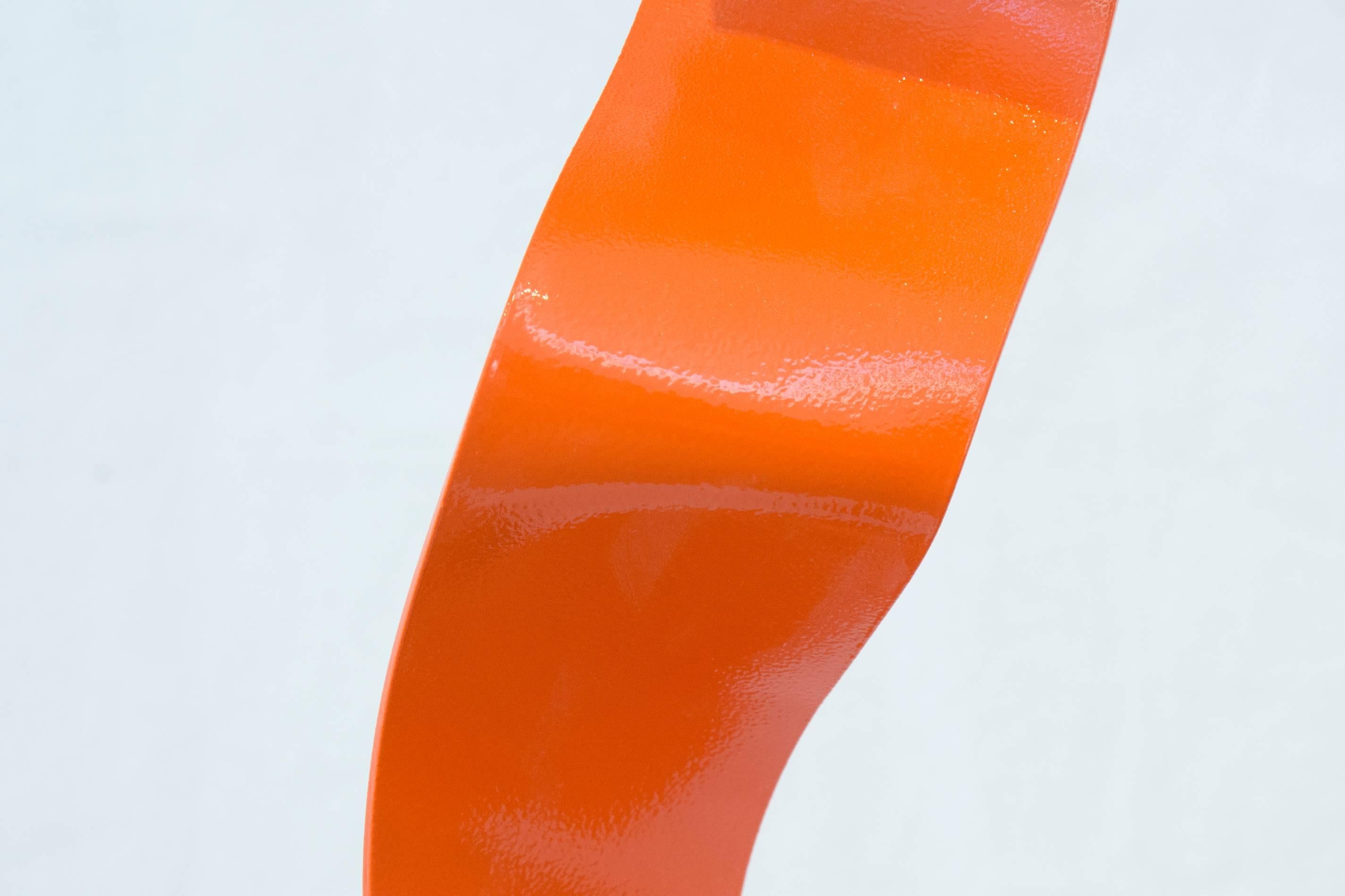 A ribbon of glossy, bright orange steel unwinds gracefully in mid air and bounces off a square, black wooden base with a dynamic flourish. The seeming effortlessness of this movement is belied by the physical process of making the sculpture. The