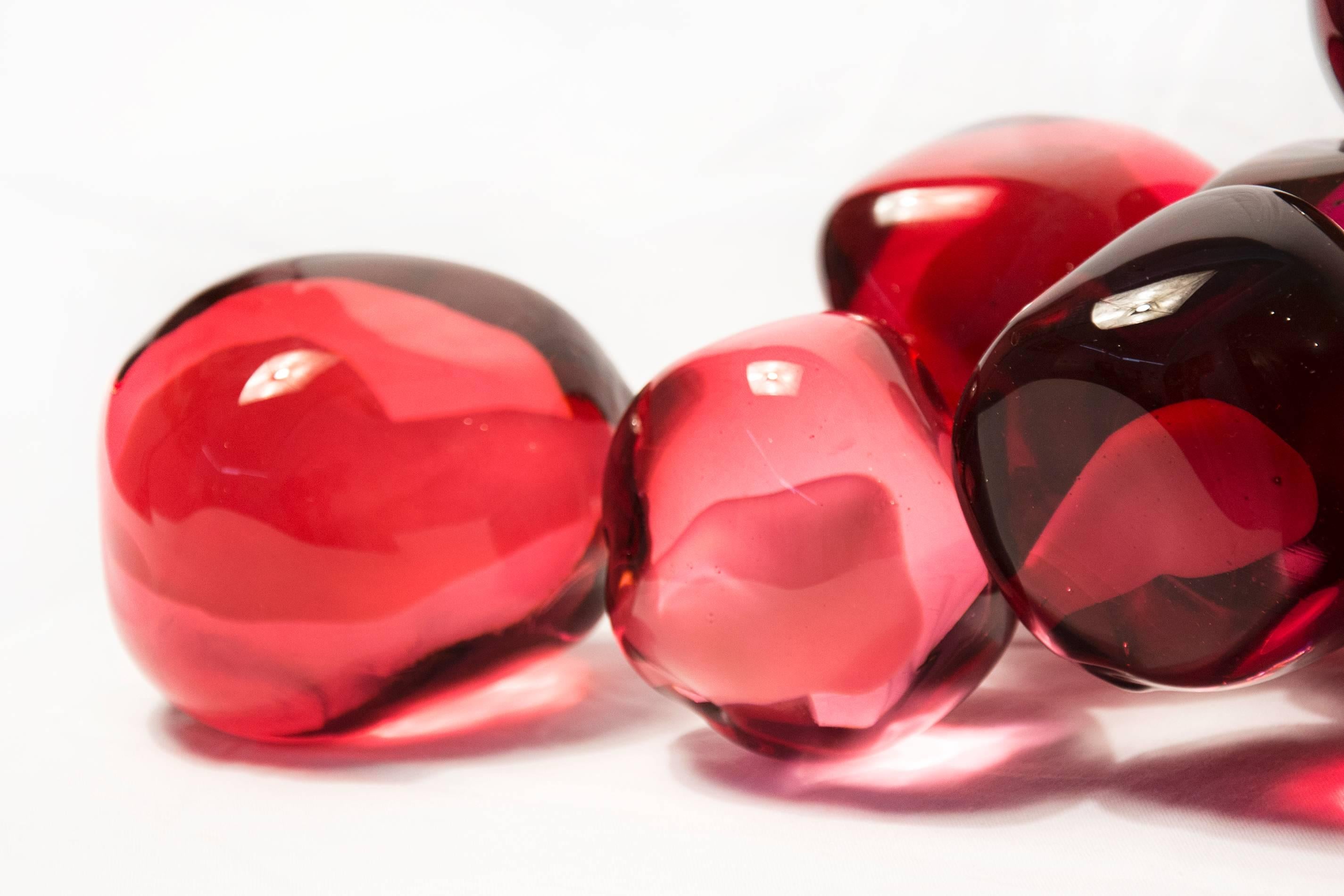 Clustered and sensuous oblong forms in translucent red glass have a smooth polished surface like the fruit that surrounds pomegranate seeds. A single seed sits next to the cluster. The pomegranate represents abundance, fortune and fertility. 

