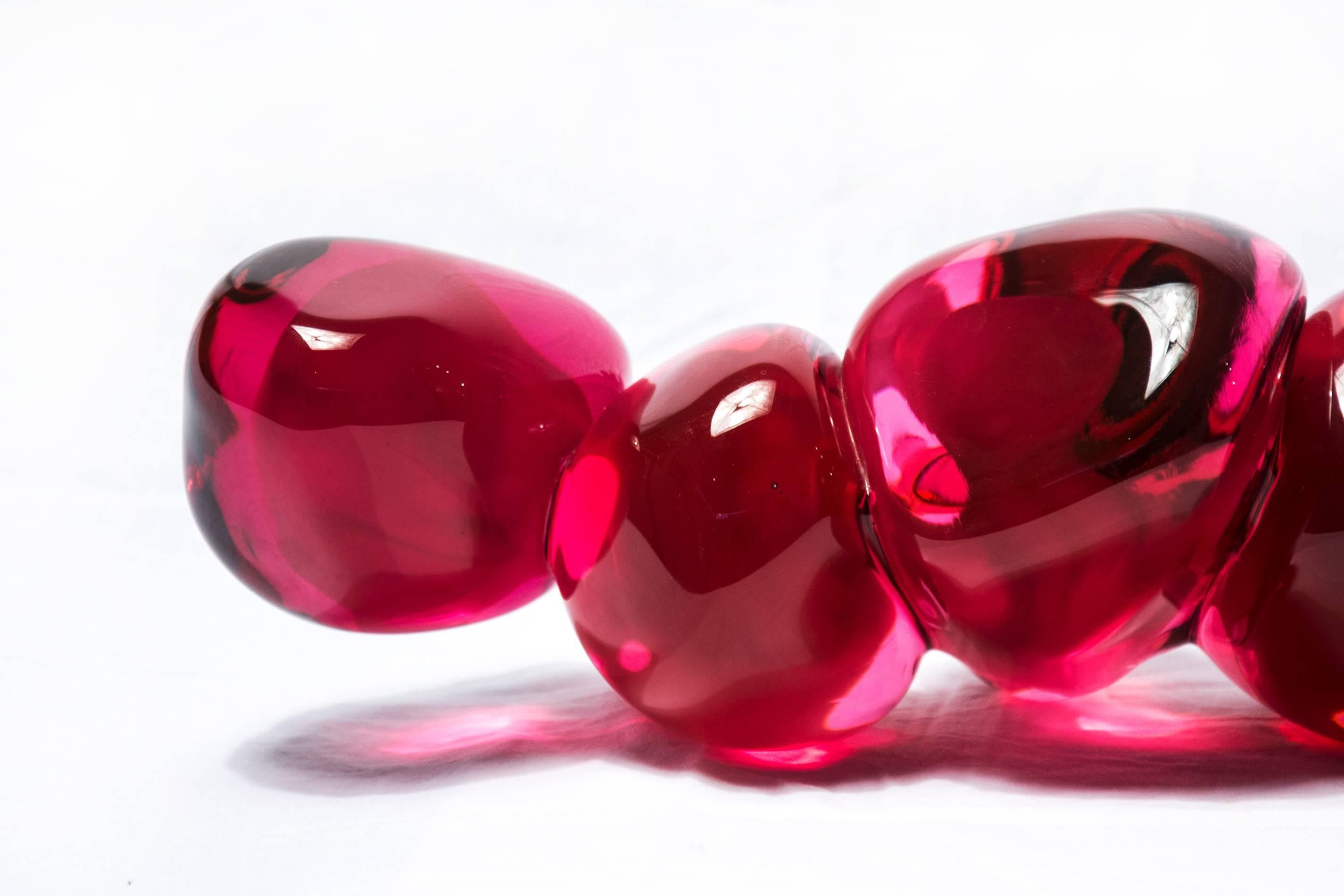 Clustered and sensuous oblong forms in translucent red glass have a smooth polished surface like the fruit that surrounds pomegranate seeds. A single seed sits next to the cluster. The pomegranate represents abundance, fortune and fertility. 

