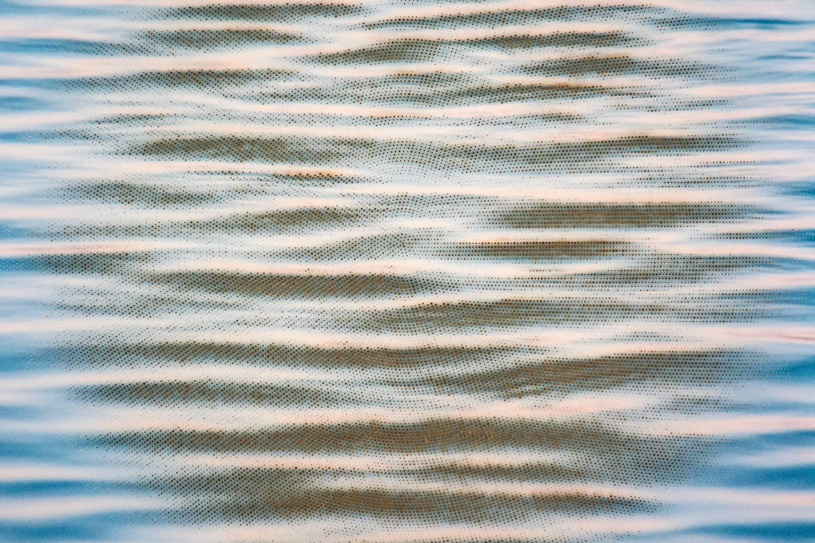 From beneath the black, light blue and off white ripples on the surface of calm water, a gold sun emerges through precise, geometric perforations in this mixed media image by Ryan Van Der Hout. Gold leaf behind a laser cut pigment print photograph