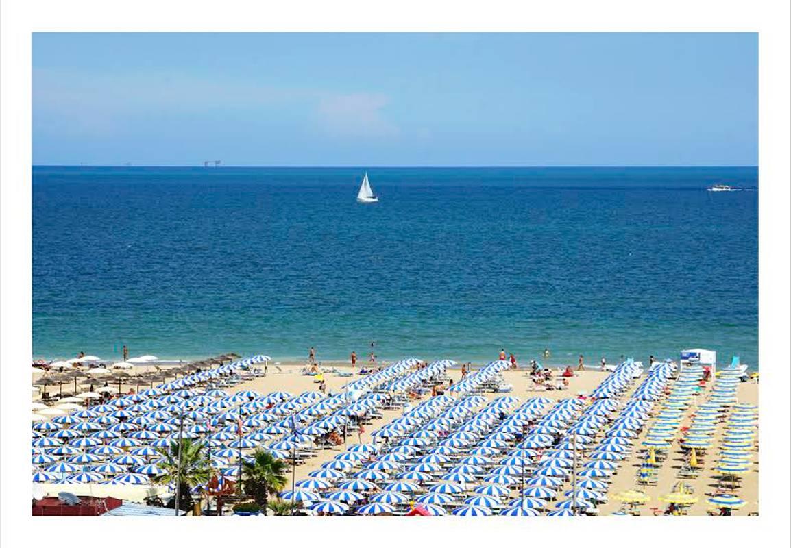 L'œuvre "Beach Umbrellas/ Rimini" de DJ Leon capture une scène de plage italienne avec un ciel et un océan d'un bleu profond, du sable jaune et un voilier blanc.  Impression C.C. numérique face montée sur plexi, 40 x 60 pouces, édition de