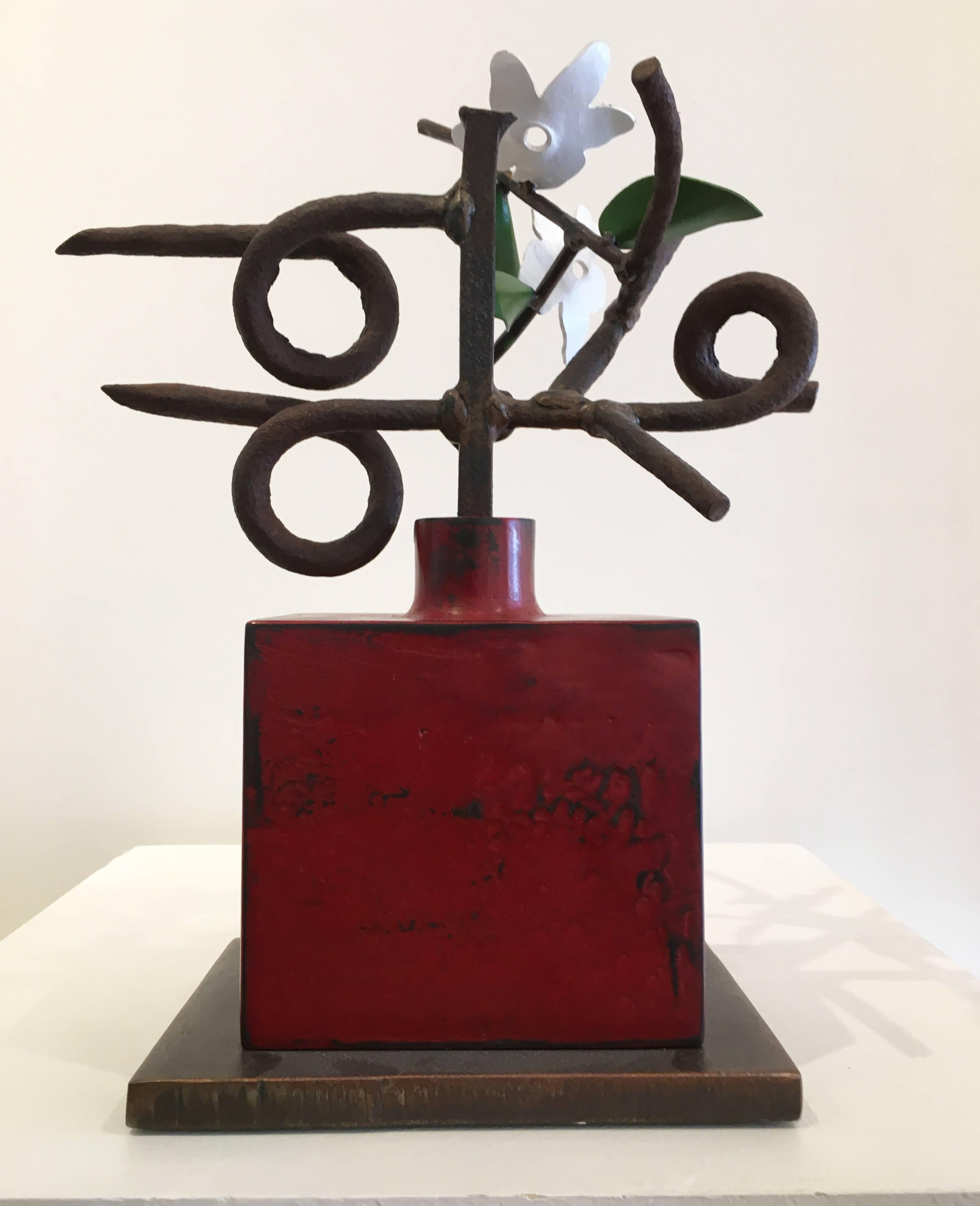 “Sanskrit” by David Kimball Anderson, 2012. Bronze, steel, and paint, 16 x 11.5 x 6 inches. This artwork features a square bronze vase in a rich red patina. Contained in the vase are two steel flowers painted in white with three green leaves. The