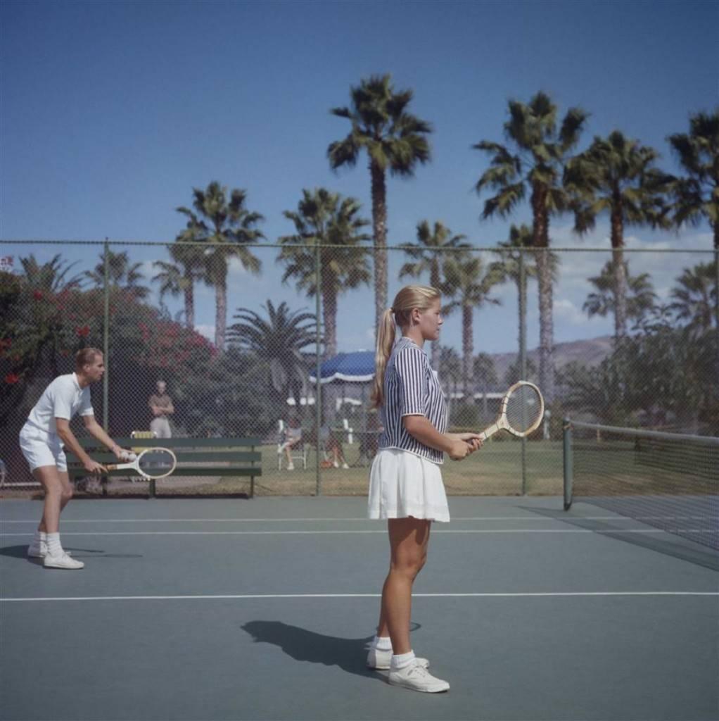 'Tennis In San Diego' by Slim Aarons

A man and a woman playing tennis on a court surrounded by palm trees, San Diego, California, October 1956.

The elegant pair dressed in tennis whites, save for the striped shirt of the young lady, enjoy a game