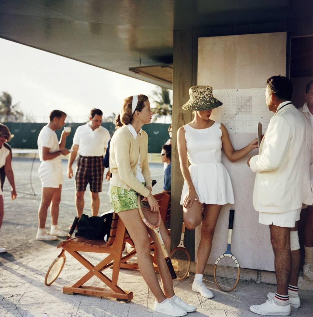 'Tennis auf den Bahamas' von Slim Aarons

Fabelhafter geht es wirklich nicht mehr.

Eine echte Neuentdeckung im Archiv: Zwei modisch gekleidete Frauen in Tennis-Outfits unterhalten sich mit einem Mann am Rande eines Tennisplatzes auf den Bahamas,