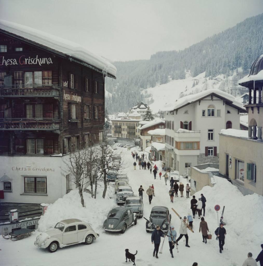 'Klosters' by Slim Aarons

Skiers pass by the Hotel Chesa Grischuna in Klosters, 1963.

Skiers walk past the hotel and along the snow covered road carrying skis on their shoulders. The road is lined with classic and vintage cars of the era and 