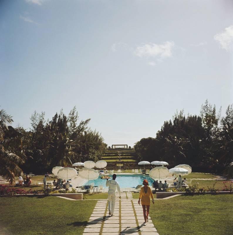 Dîner aux Bahamas 

Un serveur portant un plateau de nourriture passe devant une femme qui s'éloigne d'une piscine entourée de tables, chacune avec un parasol pour ombrager les convives, aux Bahamas, vers 1957.

Magnifique estampe mesurant un très