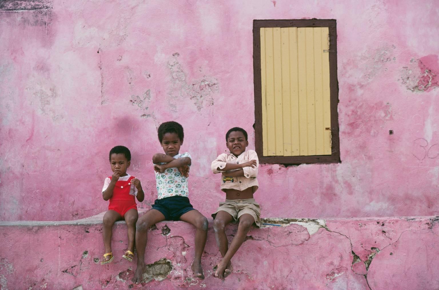 'Curacao Children' by Slim Aarons

Three local children sitting on a low predominantly pink coloured wall with a yellow shuttered window behind them, with cheeky smiles and expressions - Curacao, Netherlands Antilles, January 1979. 

A relatively