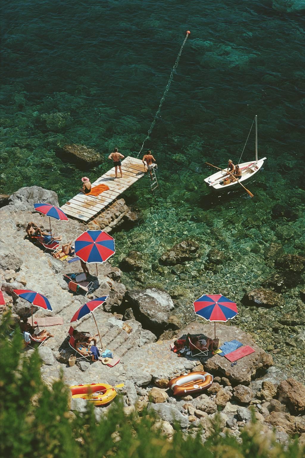 'Porto Ercole' Italy  (Slim Aarons Estate Edition)

A jetty juts out from the rocky shoreline at the Hotel Il Pellicano in Porto Ercole, Tuscany, August 1973.

Beautiful greens of the sea water contrast with the red and blue parasols, gorgeous