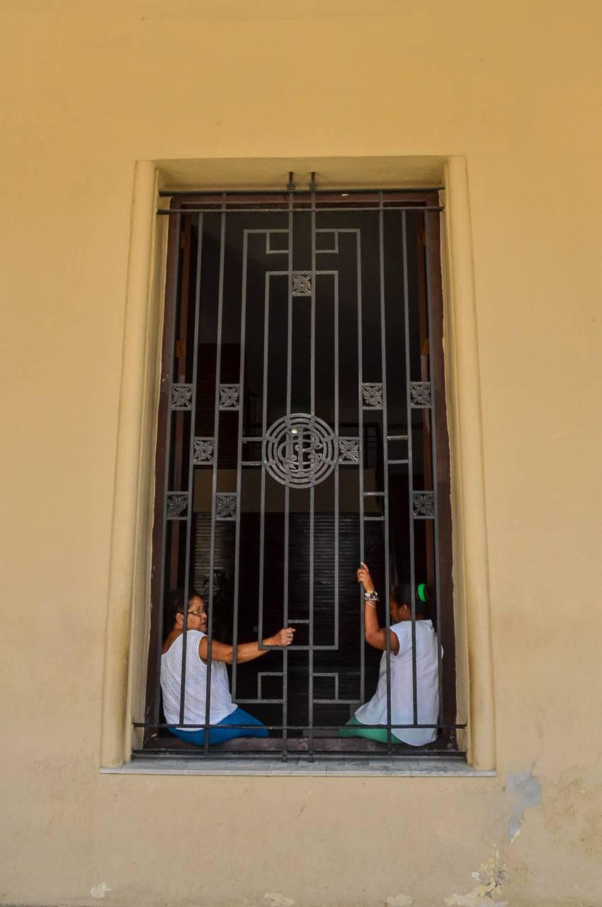 The two women in this fine art photograph lean against the wrought iron covering the double sized window, reminiscent of Andalusian courtyard windows, but in this case, a scene from Havana, Cuba.. The wrought iron design, with elements of art deco,