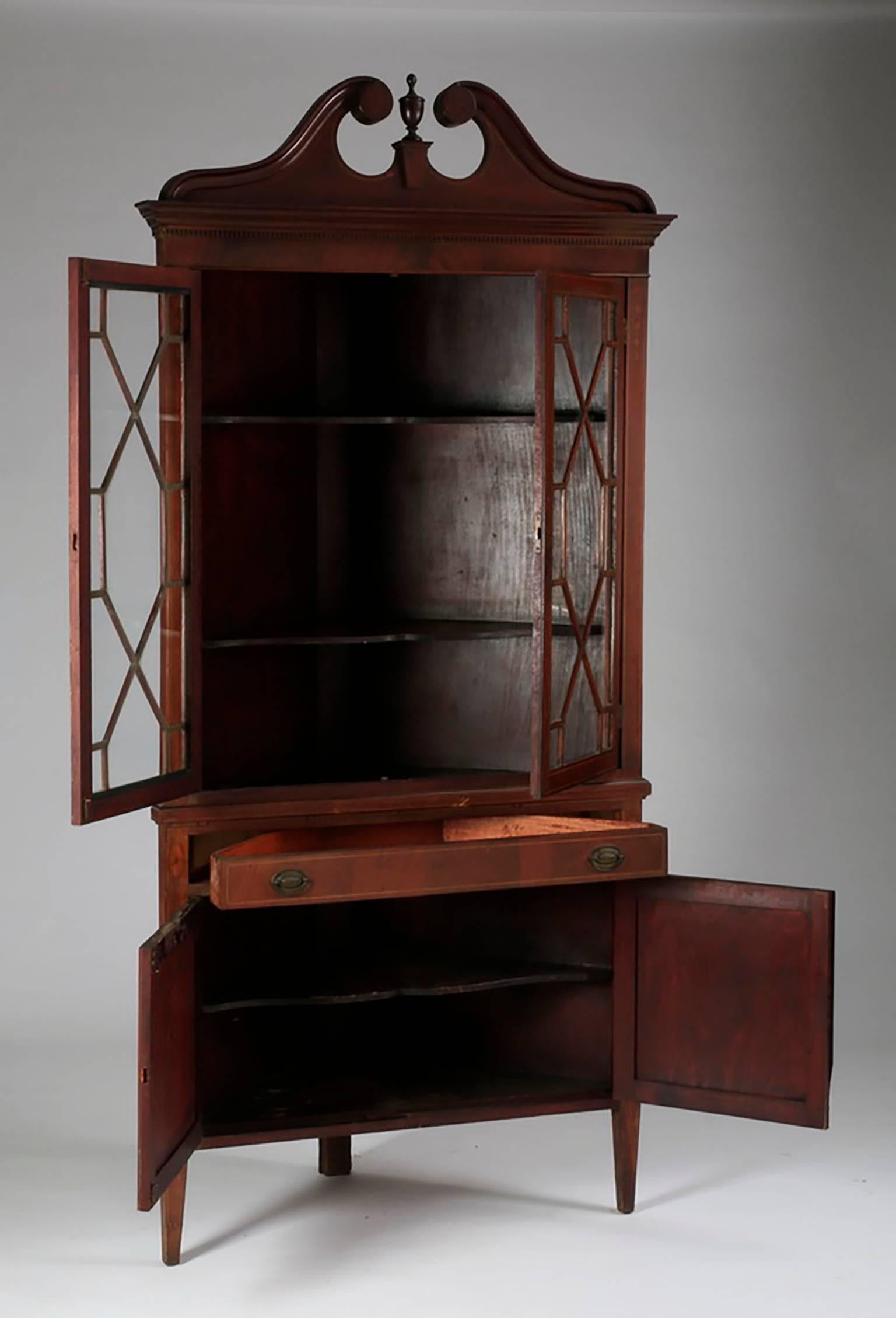 Antique American Georgian style mahogany double door corner cabinet. It has an arch pediment with marquetry inlaid decoration above the stepped rectangular top surmounting two glass doors that open to reveal three display shelves. There is one long