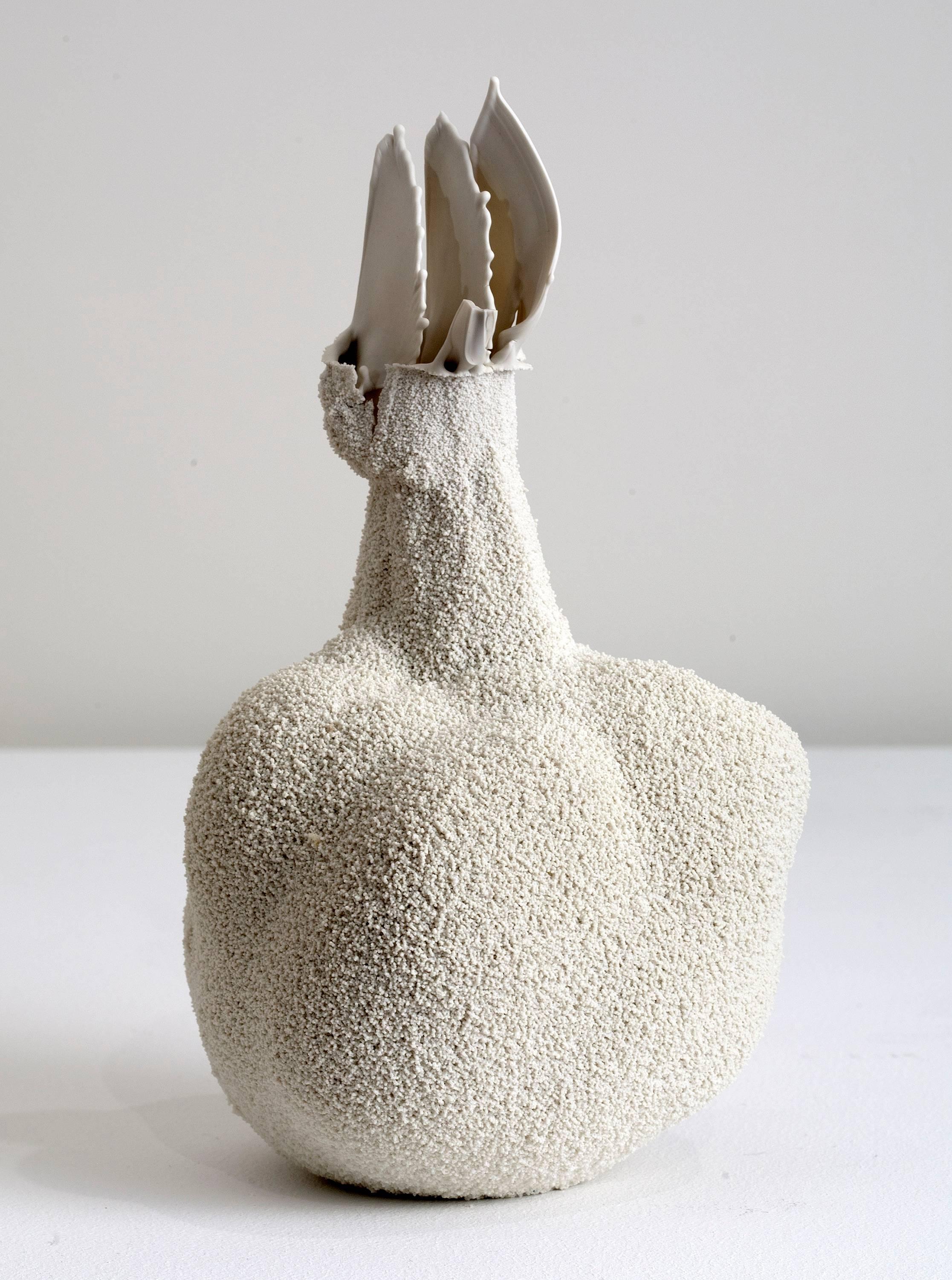 This is a small white ceramic vase by London-based Israeli artist Michal Fargo. She pours liquid porcelain into a hollowed block of foam, allowing it to settle into the pores. The vessel's final shape is determined by the material rather than the