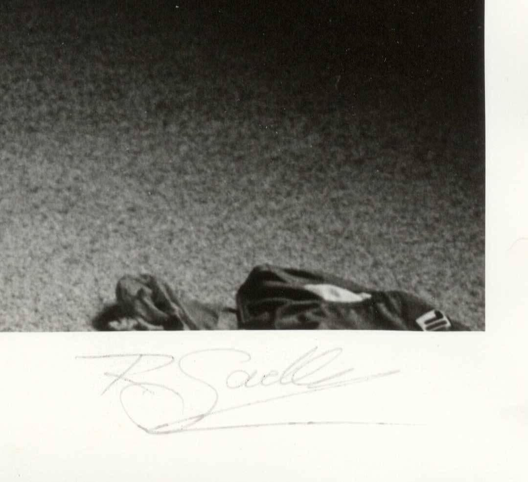 The image shows a full-frontal photographic portrait of a young nude male gazing out of a window. Items of clothing are seen lying on the floor.  Richard Sadler created this silver gelatin photograph in 1981 in Arles, France while on a trip with