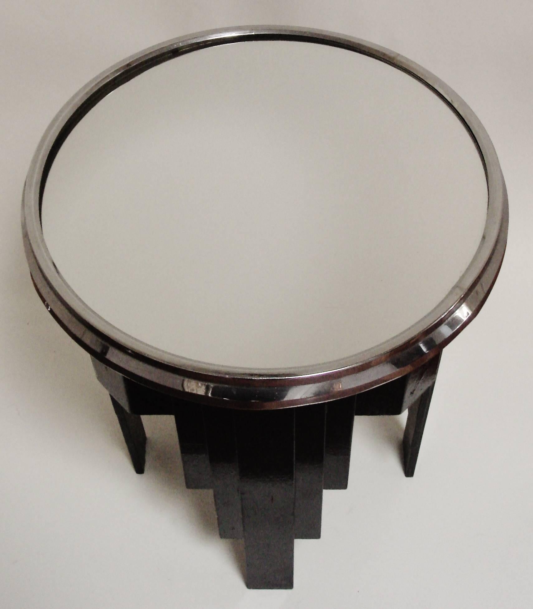 This English Art Deco circular wine table has a layered reverse ziggurat four-legged frame in wood that has been lacquered in black enamel. The mirror top is banded with a chrome gallery and the underside of the table carries a brass plaque stating