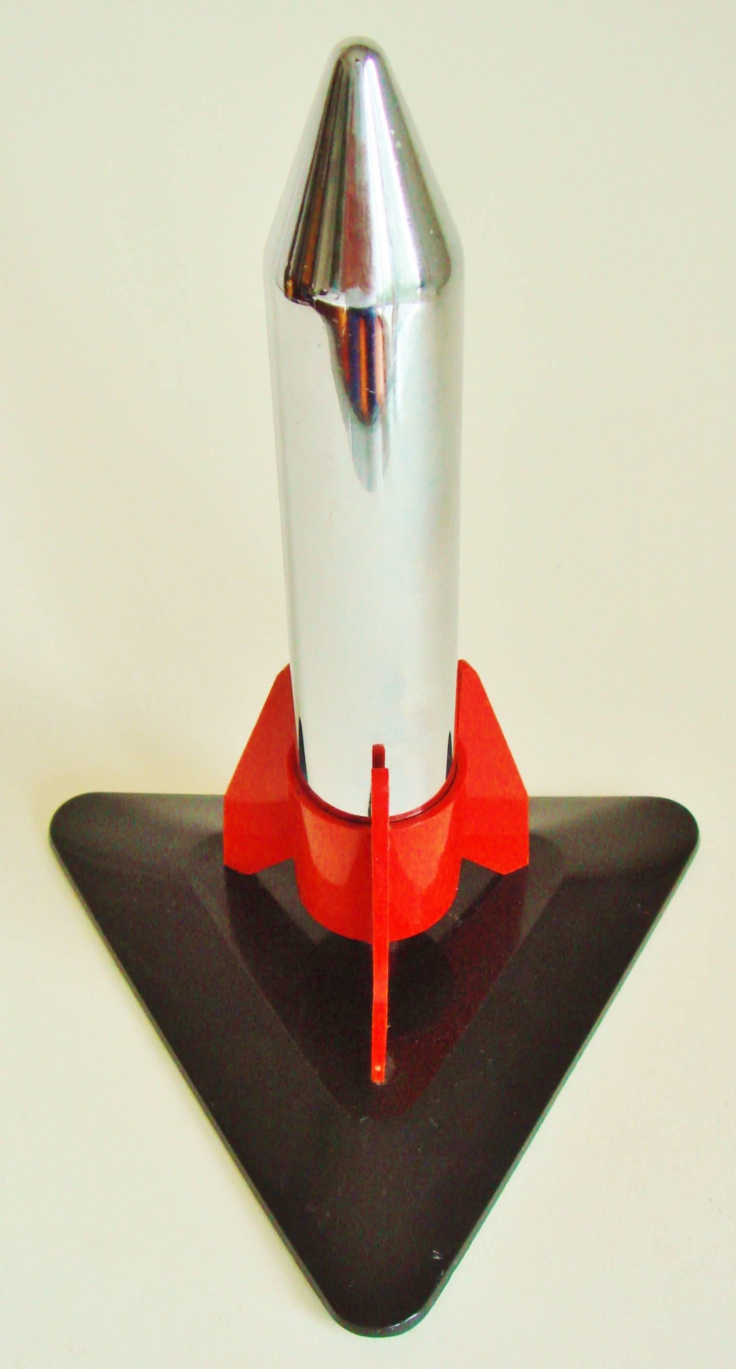 This Japanese made Mid-Century Modern wheel and flint table lighter features a chrome plated rocket body with red plastic fins and stands on a black plastic triangular base. It would appear this lighter has never been used as both the wick and