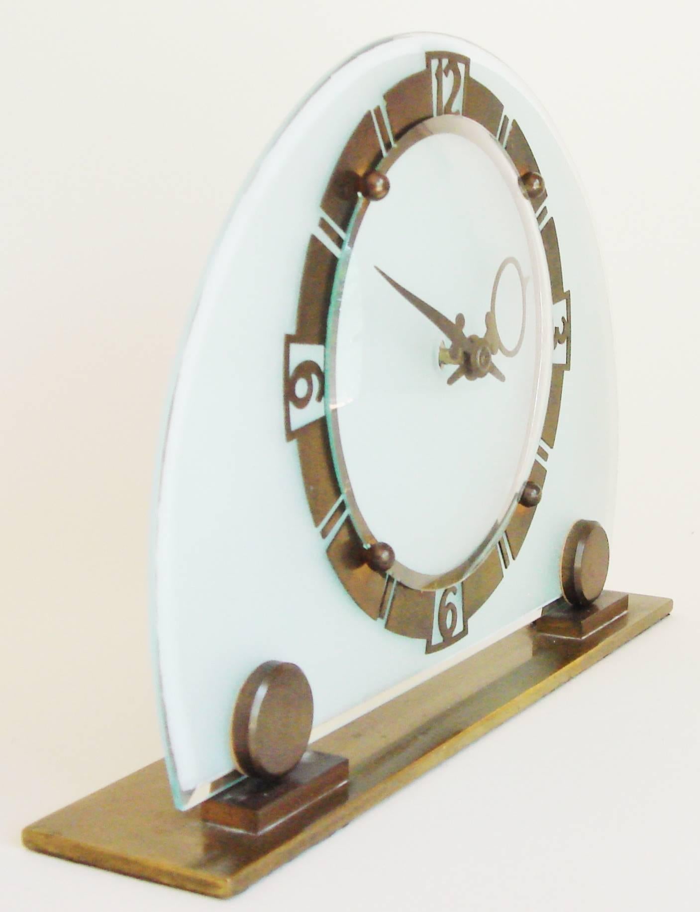 This Classic English Art Deco mechanical table/shelf clock features a frosted and beveled demilune of glass with a highly stylized cut brass chapter ring and extreme Art Deco hands. The face is covered with a convex glass that is anchored by four