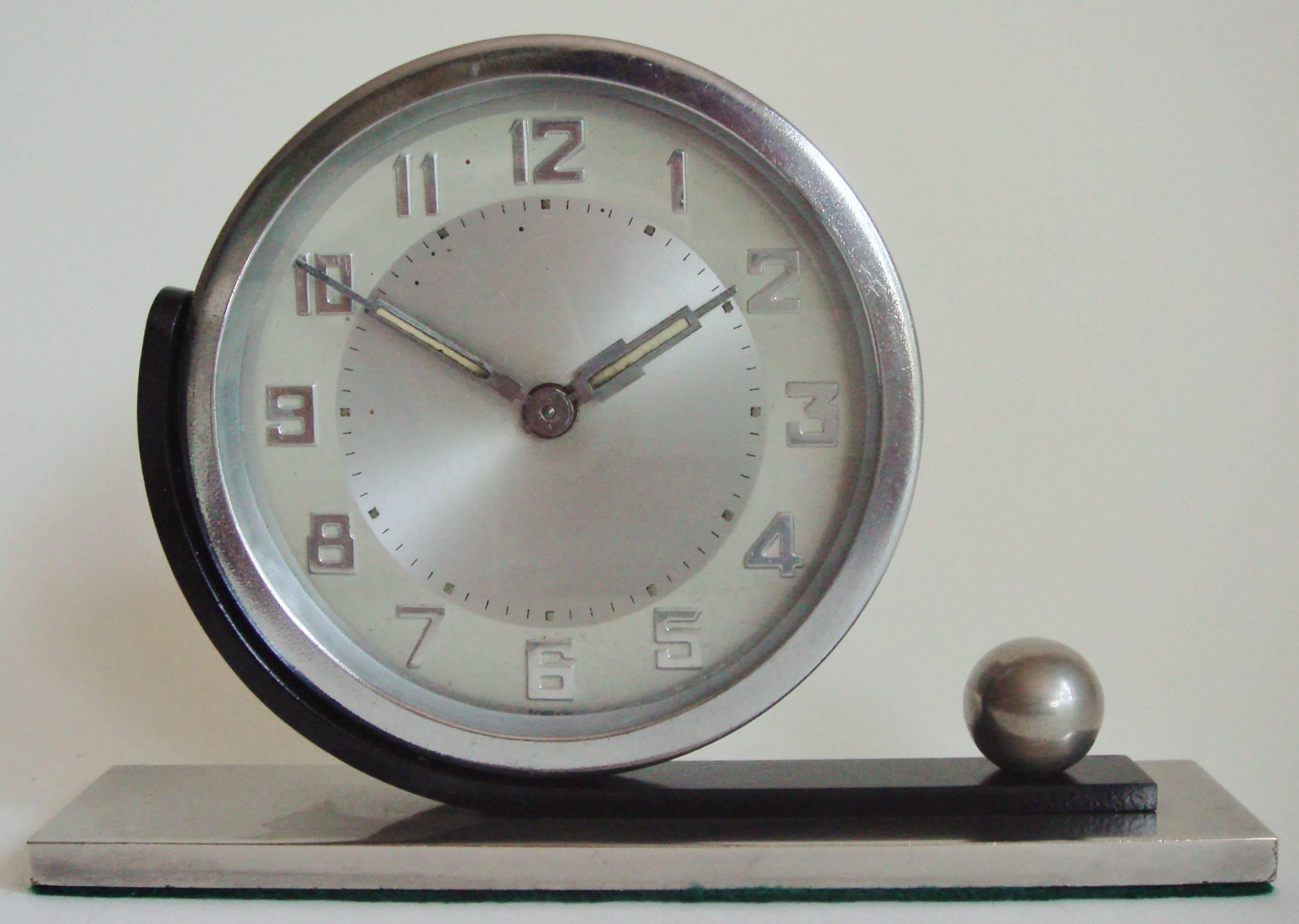 This German Art Deco asymmetrical table clock features a round clock with a chrome bezel held on a curved blackened steel arm that is anchored at one end by a brushed steel sphere. This is mounted to a solid brushed steel base with a green felt