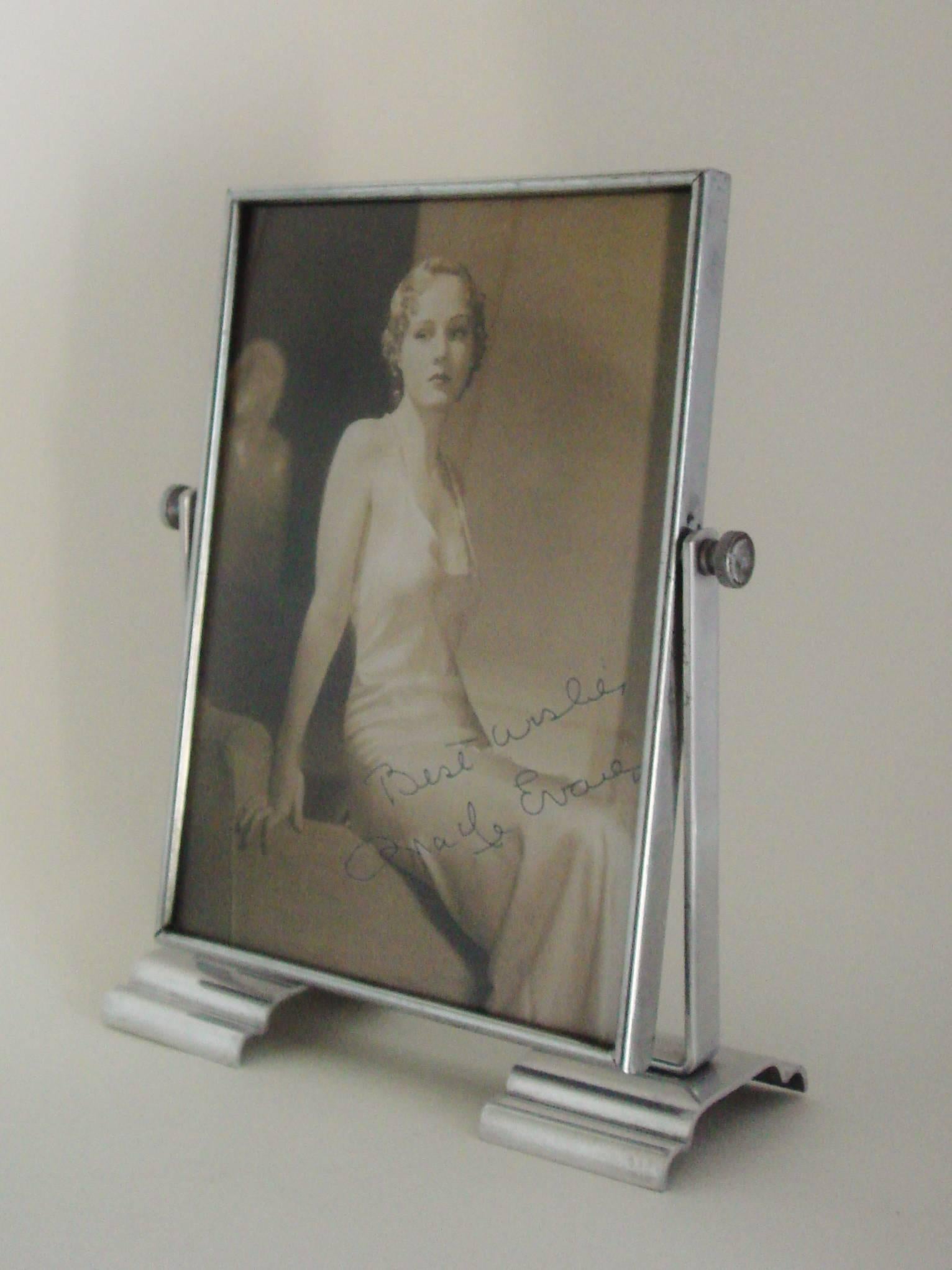 This English Art Deco adjustable photo frame features a polished aluminum base and bracket that holds a chrome-plated picture frame (8 in x 6 in image area). It stands on two polished chrome wave-form feet and has chrome bosses on either side with