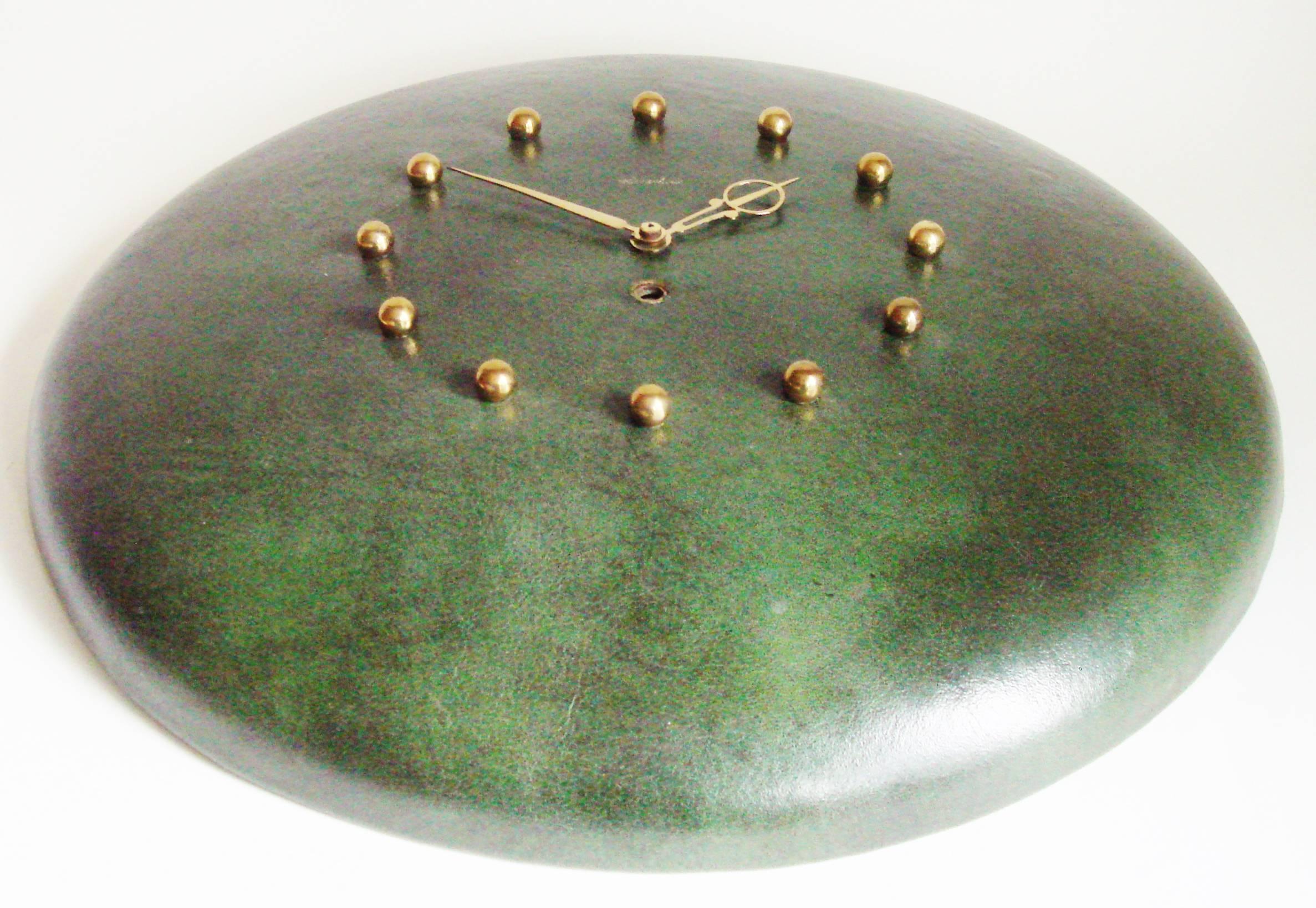 This American Mid-Century Modern large mechanical eight-day wall clock is made from base metal wrapped in mottled dark green leather. The numerals are represented by small polished brass spheres and the ornate hands are in polished brass also. The