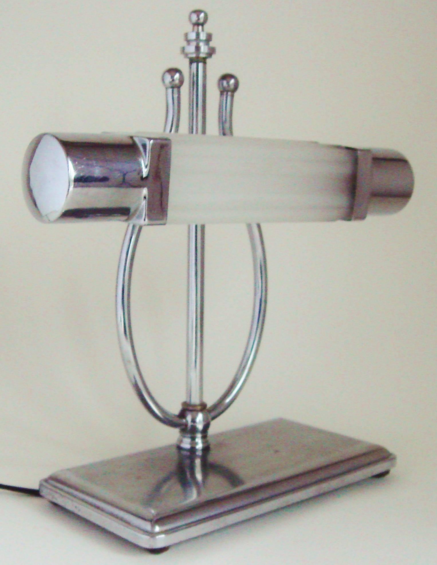 This American Art Deco piano lamp features a ribbed rectangular frosted glass shade with chrome caps at either end. This is affixed to a lyre shaped support that rises from a rectangular shaped, polished and brushed chrome base standing on four bun