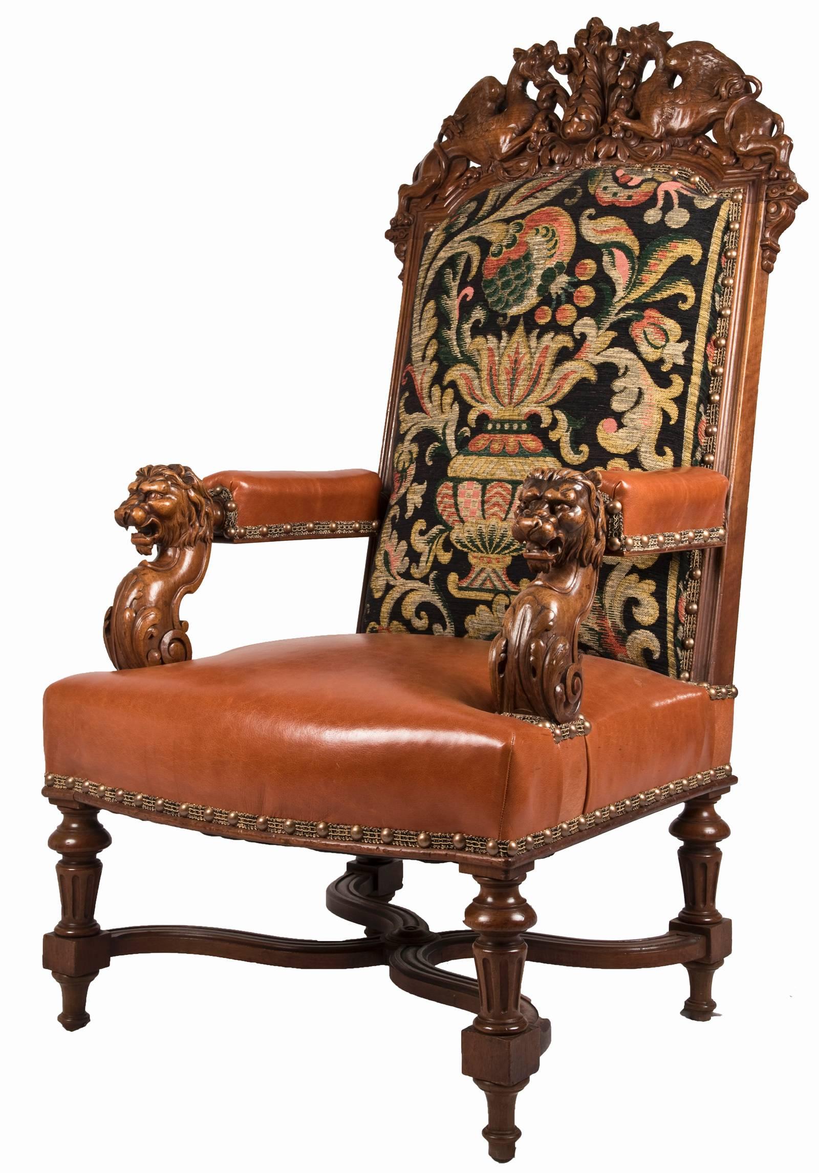 A pair of Louis XIV style fauteuil walnut chairs with a carved griffin crest, gros-point upholstered back and leather seat and arm rests with spaced brass stud trim, terminating in carved lion heads, and sitting on turned legs with an x-frame