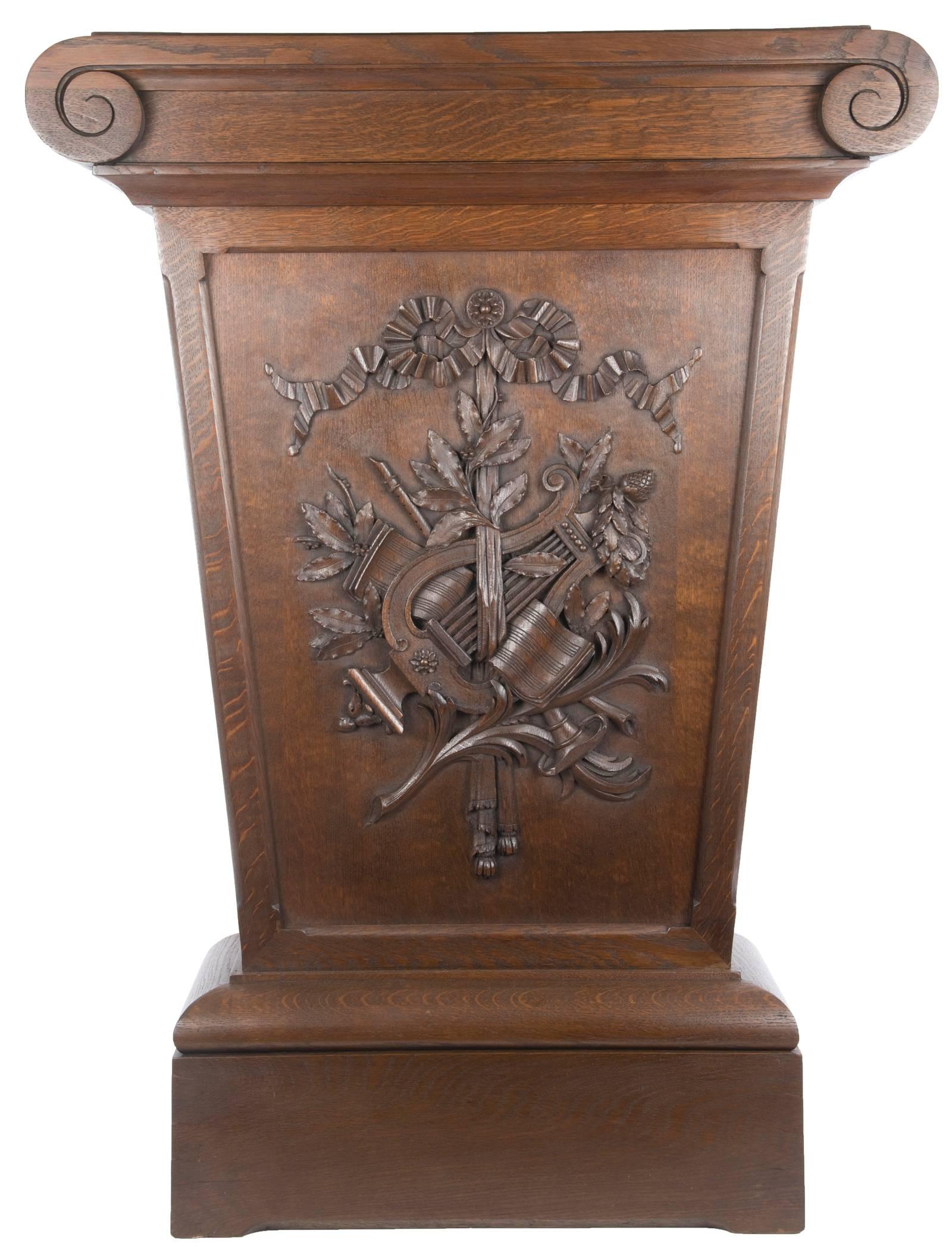 An oak pedestal stand with carved and turned scroll top, tapering form and raised on simple plinth base. Hanging from a carved rosette and tied stylized ribbon, the front panel is decorated with a carved applique of a harp and music sheets, clarinet